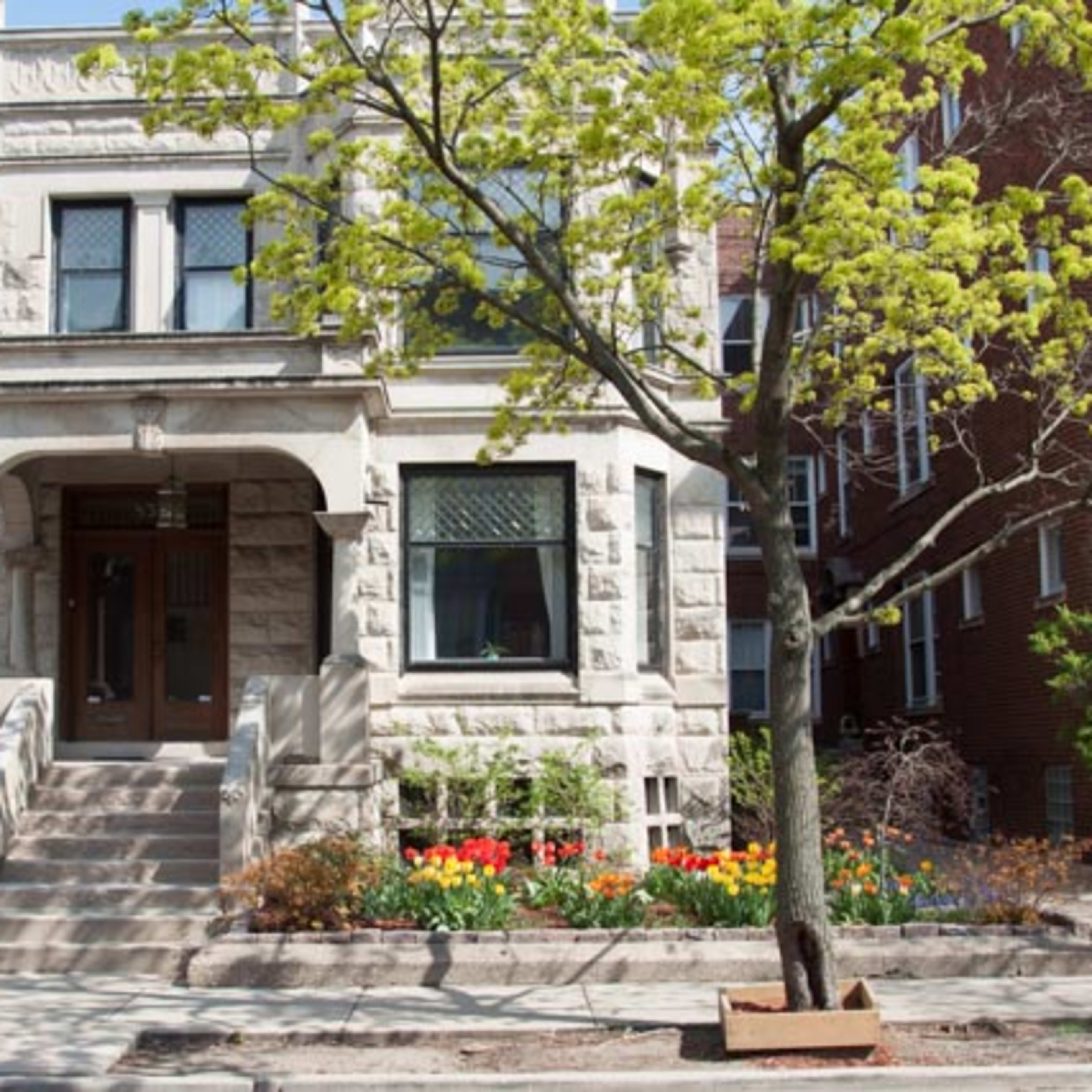 A three-story stone-faced residential building with a front porch is surrounded by flower beds and a small tree.