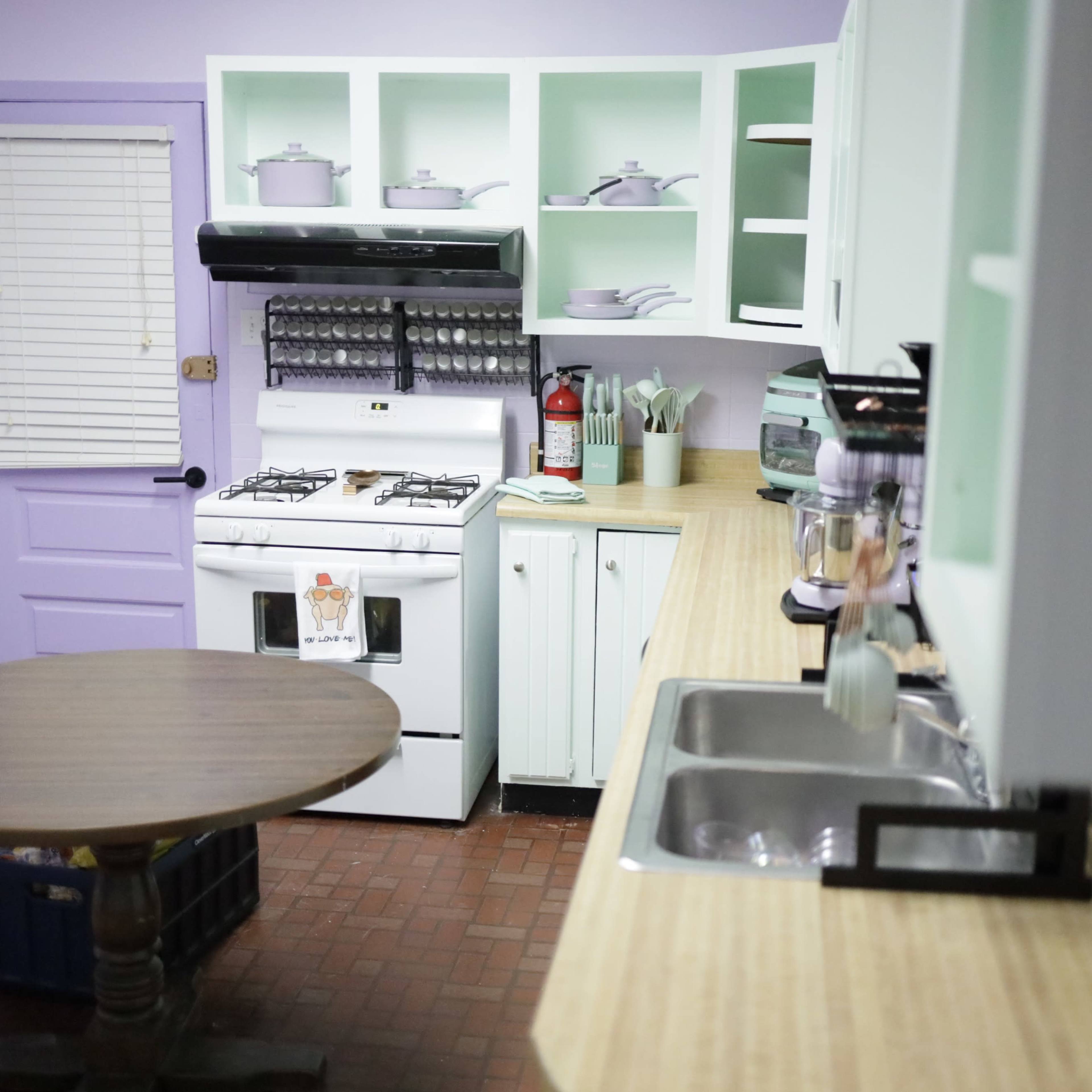 The kitchen features a teal and white color scheme with a wooden table, an oven, and various kitchen utensils on the countertops.