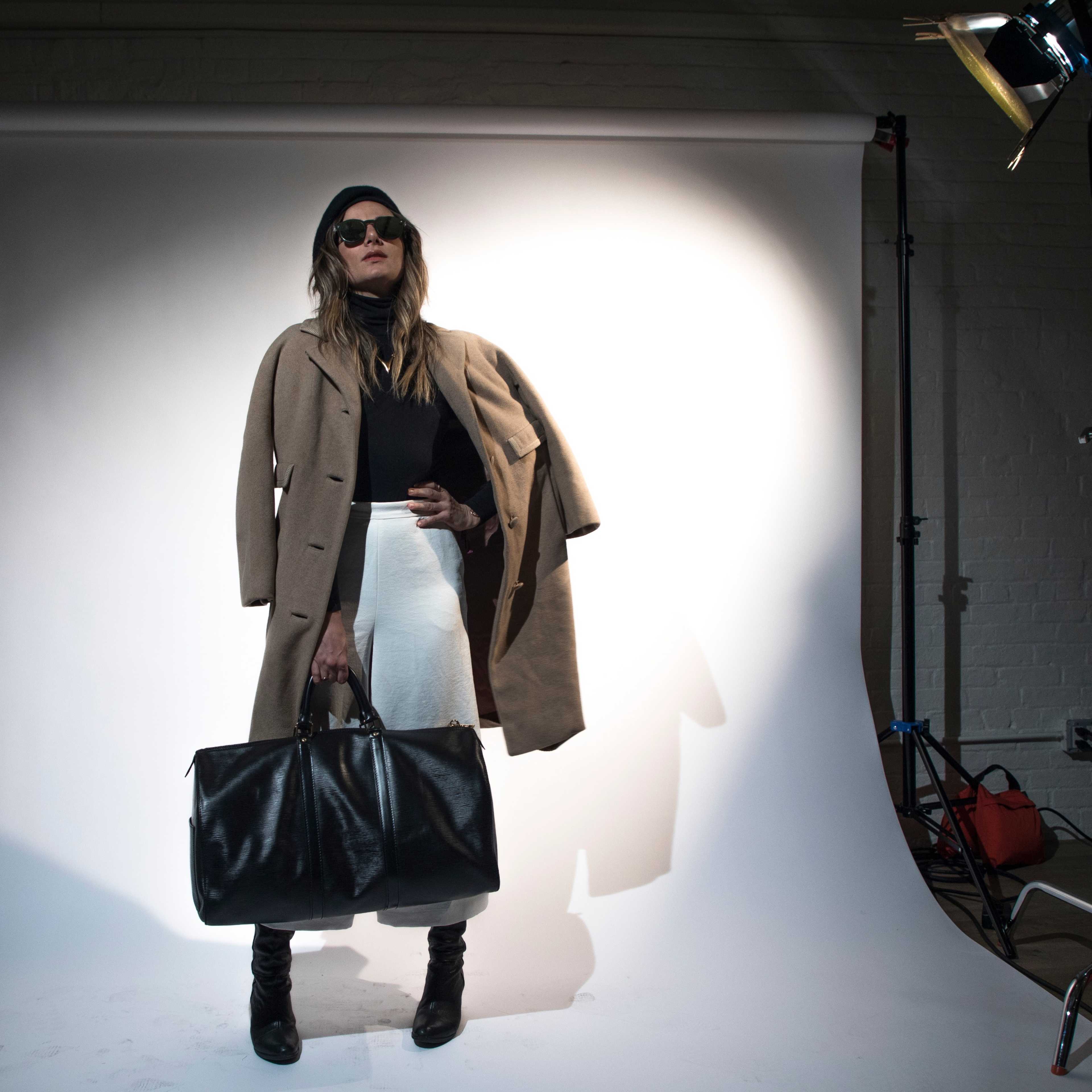 A person stands confidently in front of a white backdrop, wearing sunglasses, a long coat, and holding a large black bag.
