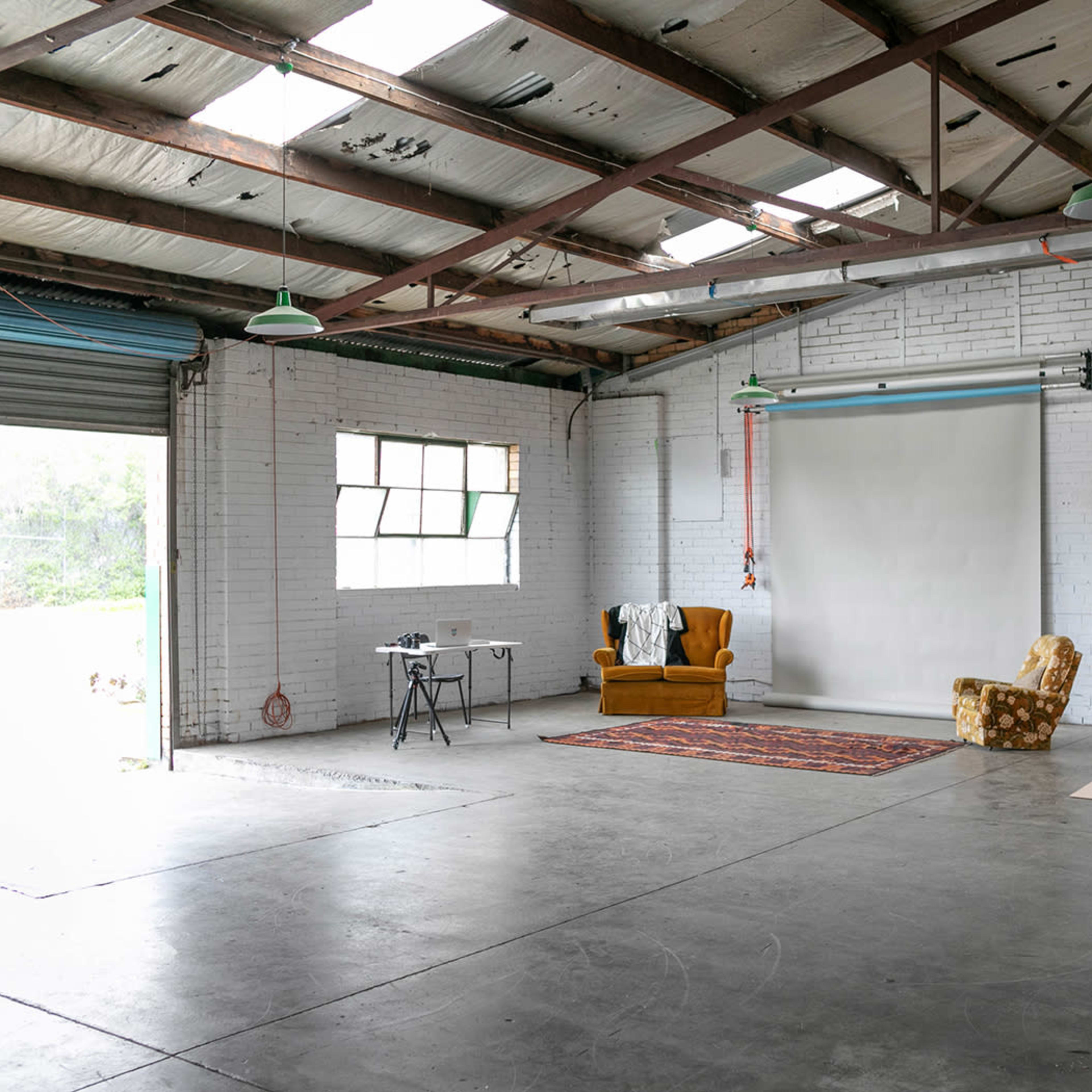 A spacious, industrial-style studio with a wooden floor, a blank backdrop, a vintage armchair, and a table with equipment set against a windowed wall.