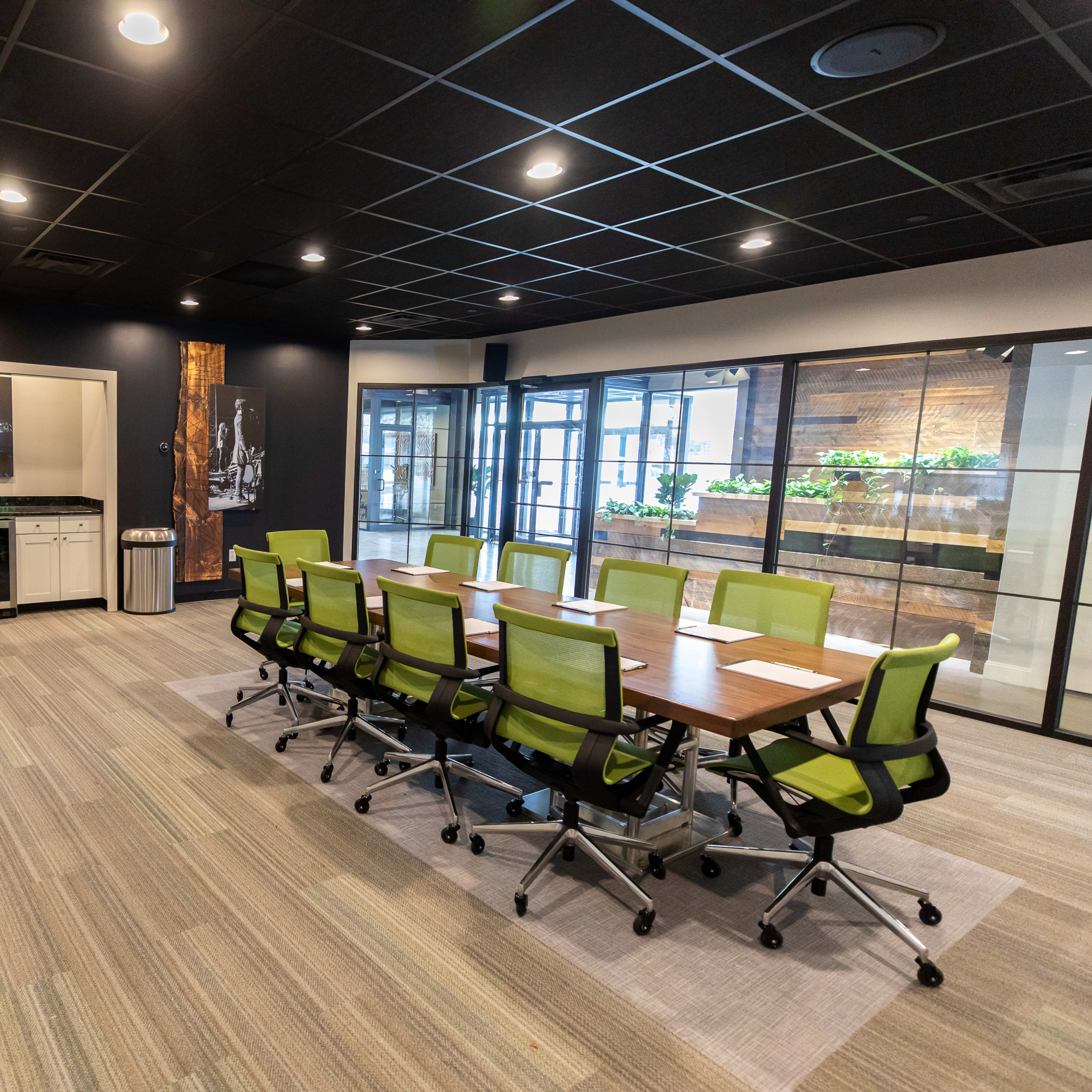 A modern conference room features a long wooden table surrounded by green rolling chairs, with a kitchenette and glass doors leading to a plant-filled area.