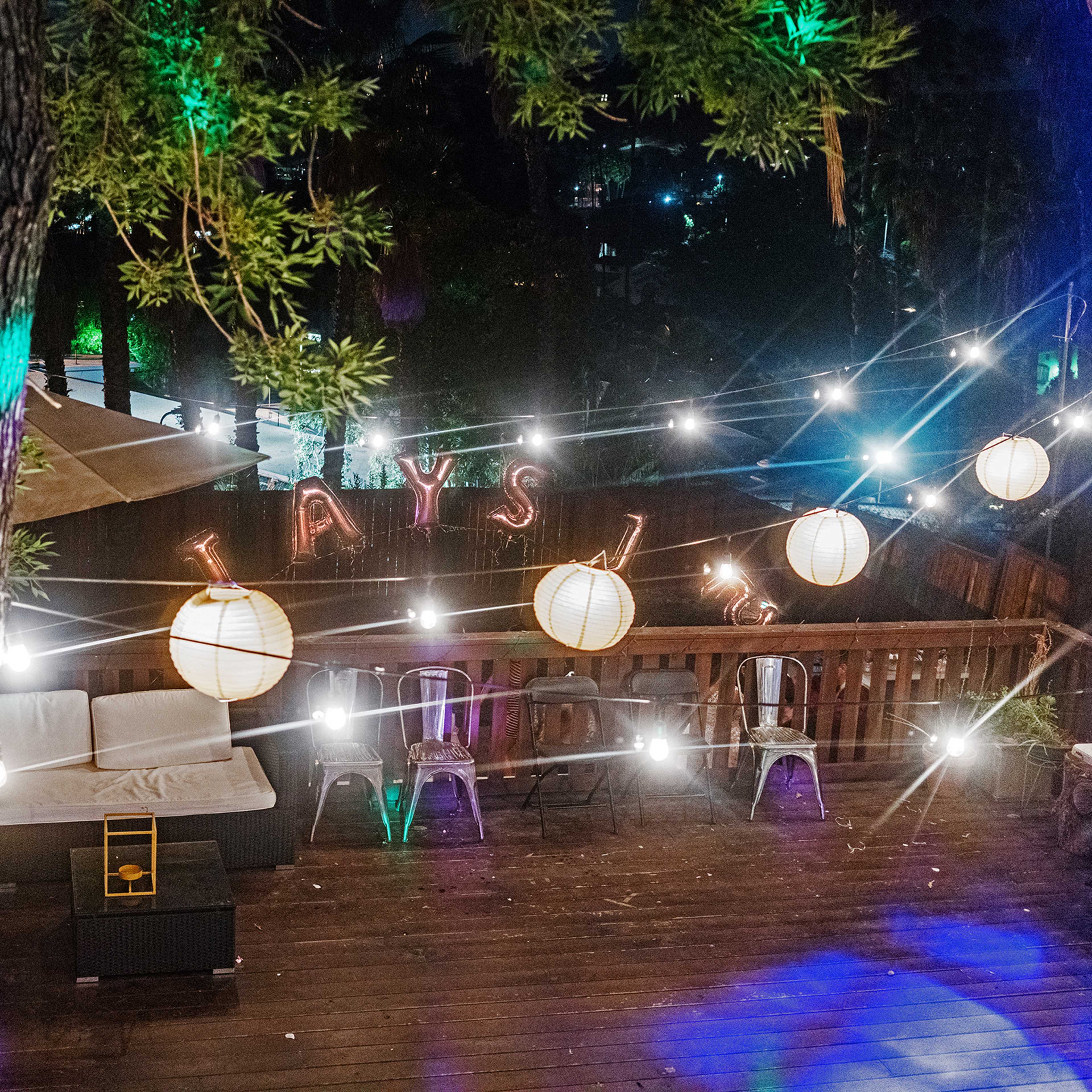 A wooden deck is illuminated by string lights and paper lanterns, featuring a seating area with couches and chairs.