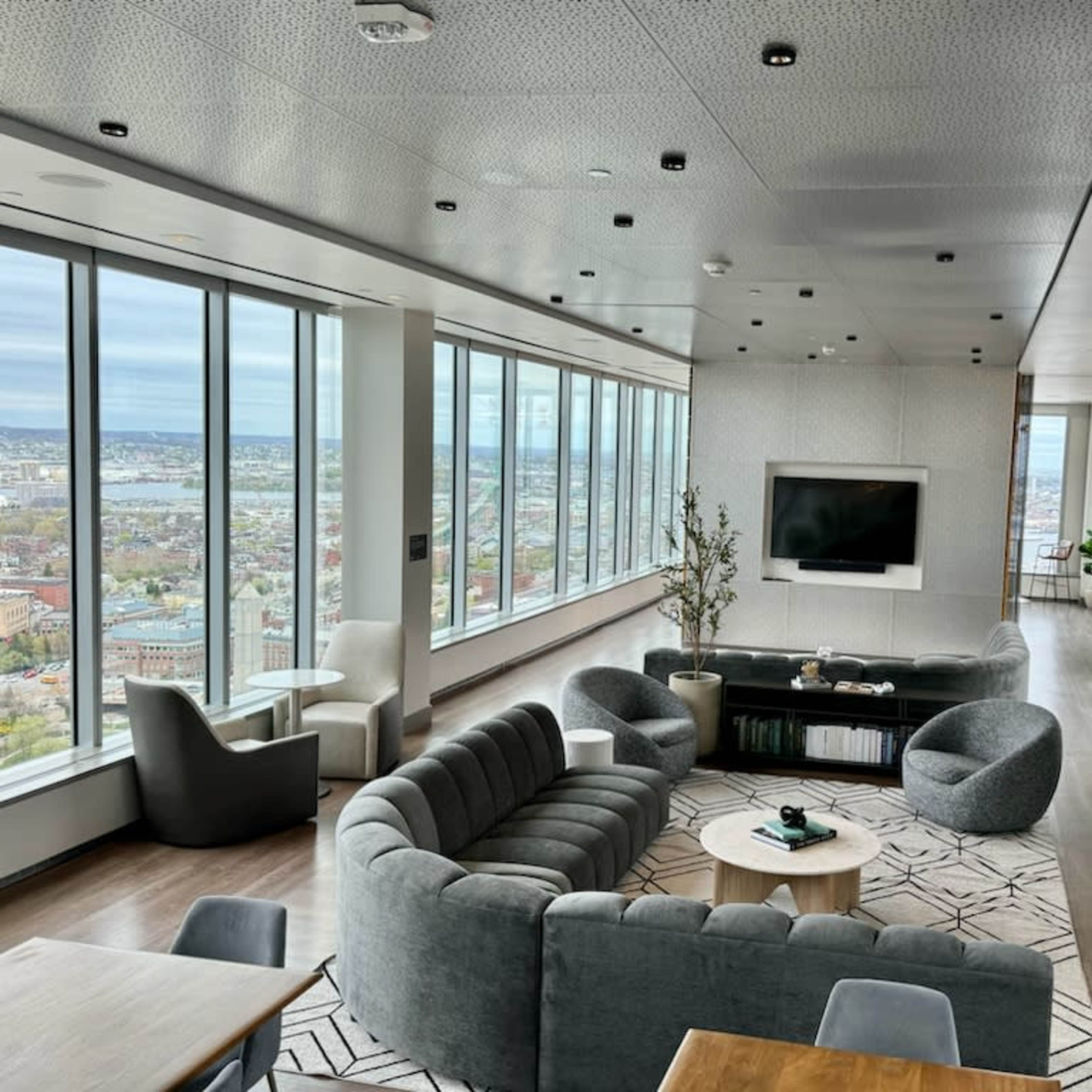 A modern lounge with large windows offering a city view, featuring gray furniture and a central coffee table.