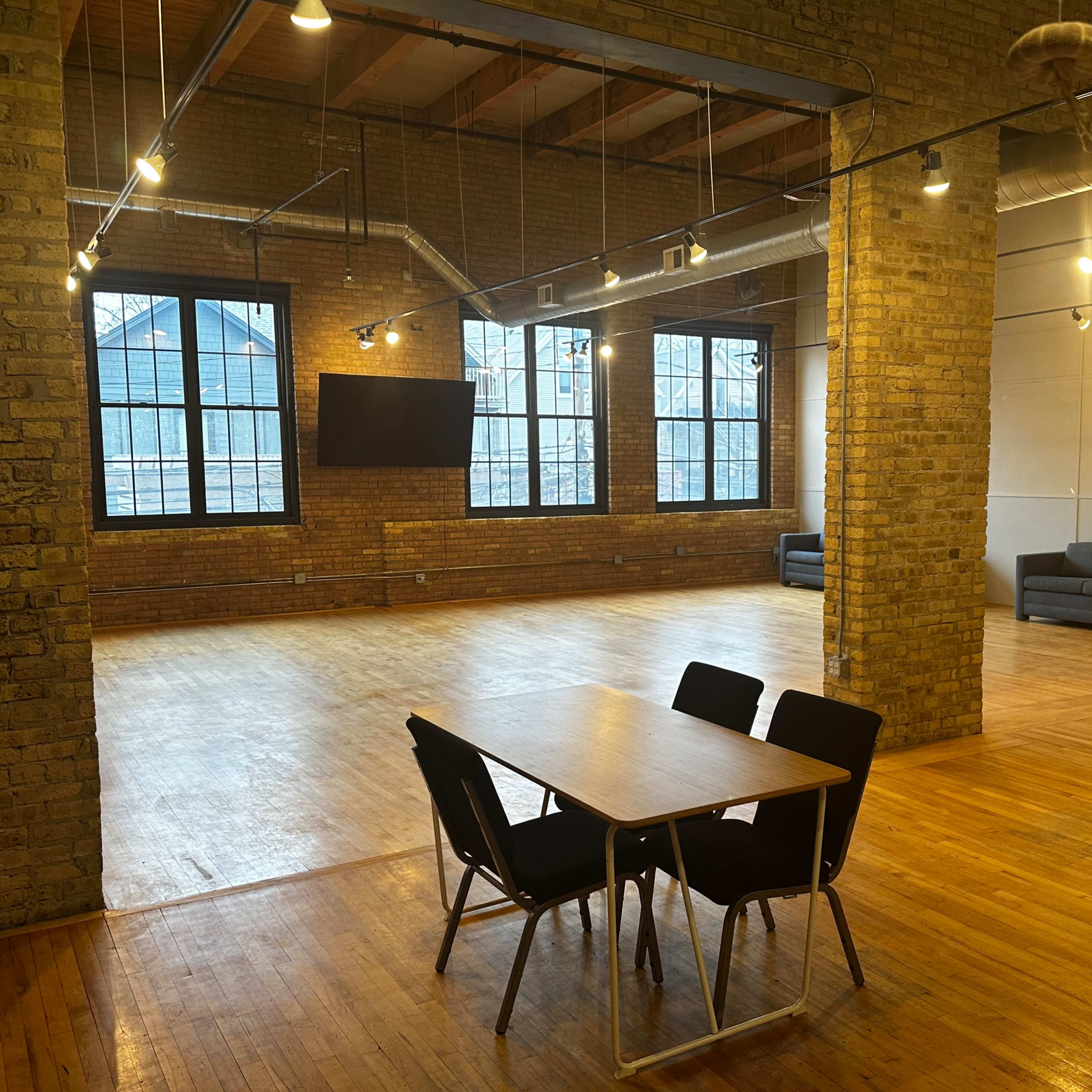 An industrial-style interior space with exposed brick walls, large windows, wooden flooring, a table with chairs, and a sofa area.