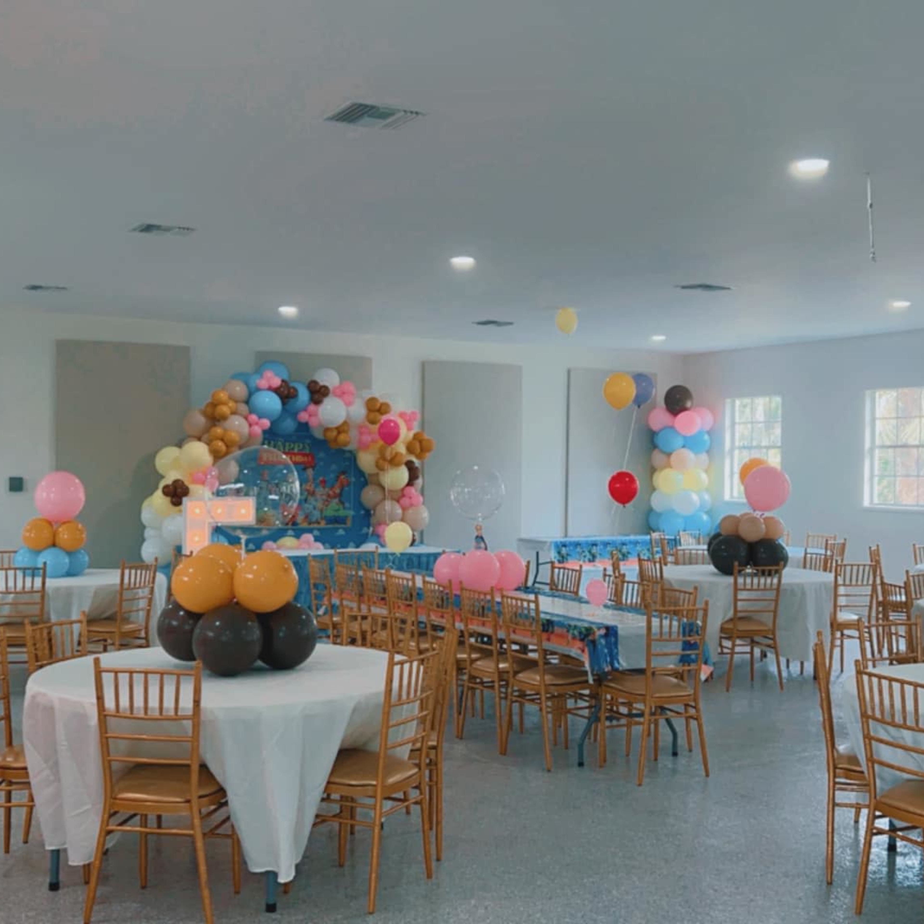 A well-decorated event space with round tables covered in white tablecloths, each adorned with balloon centerpieces, and a colorful backdrop of balloons at the front.