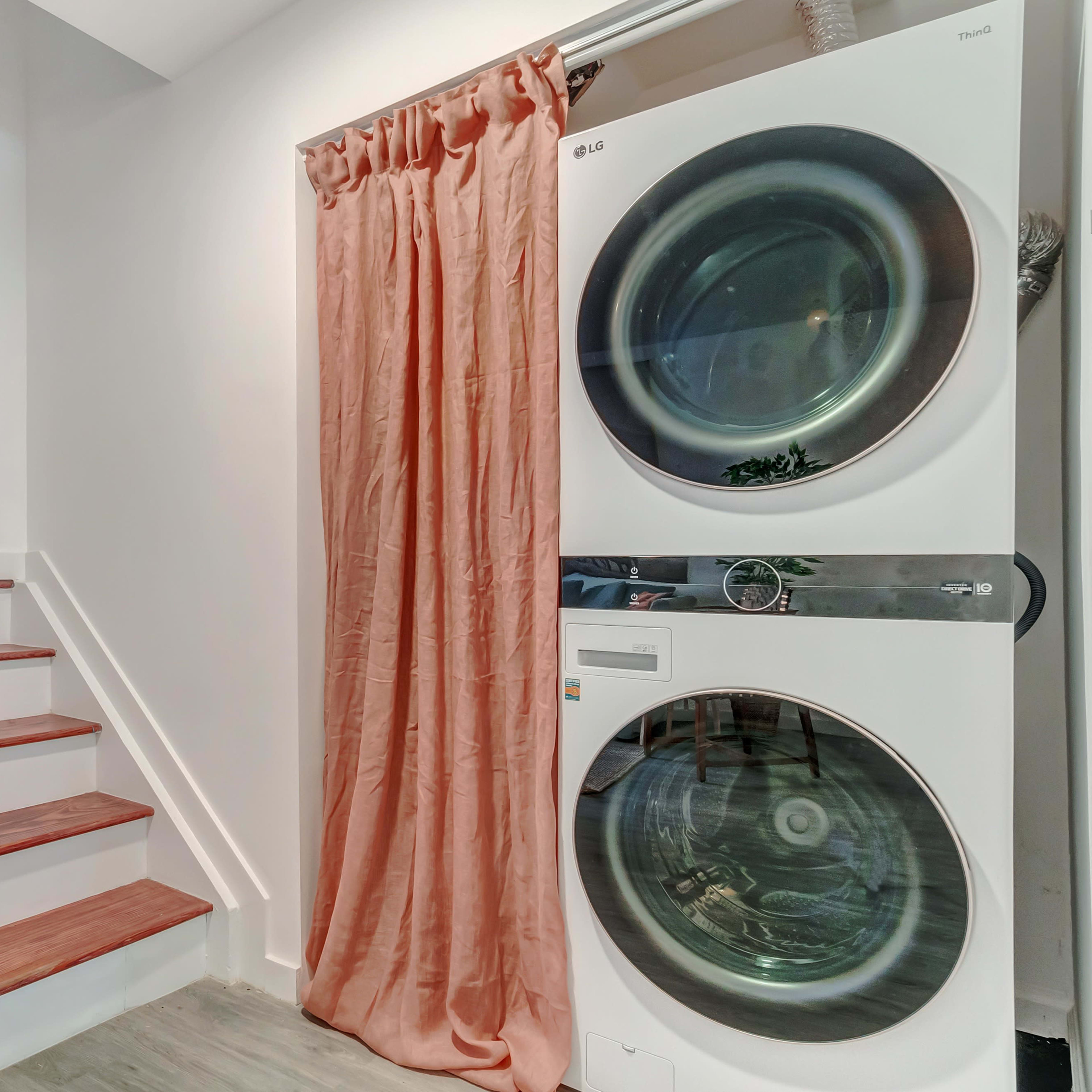A stacked washer and dryer are positioned next to a staircase, with a curtain hanging beside them.
