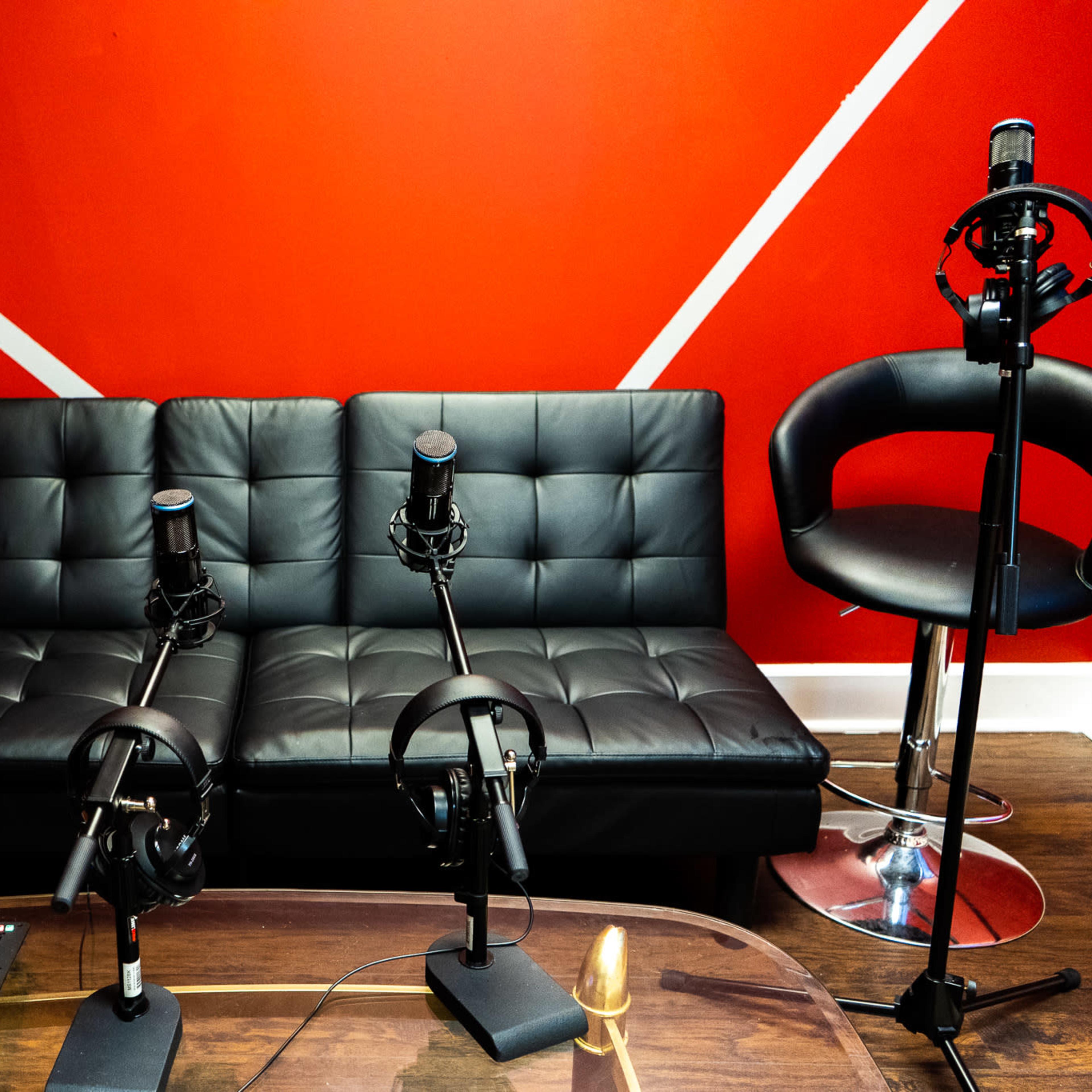 A black couch with two microphones mounted on stands in front of it, set against a red wall with white diagonal lines.
