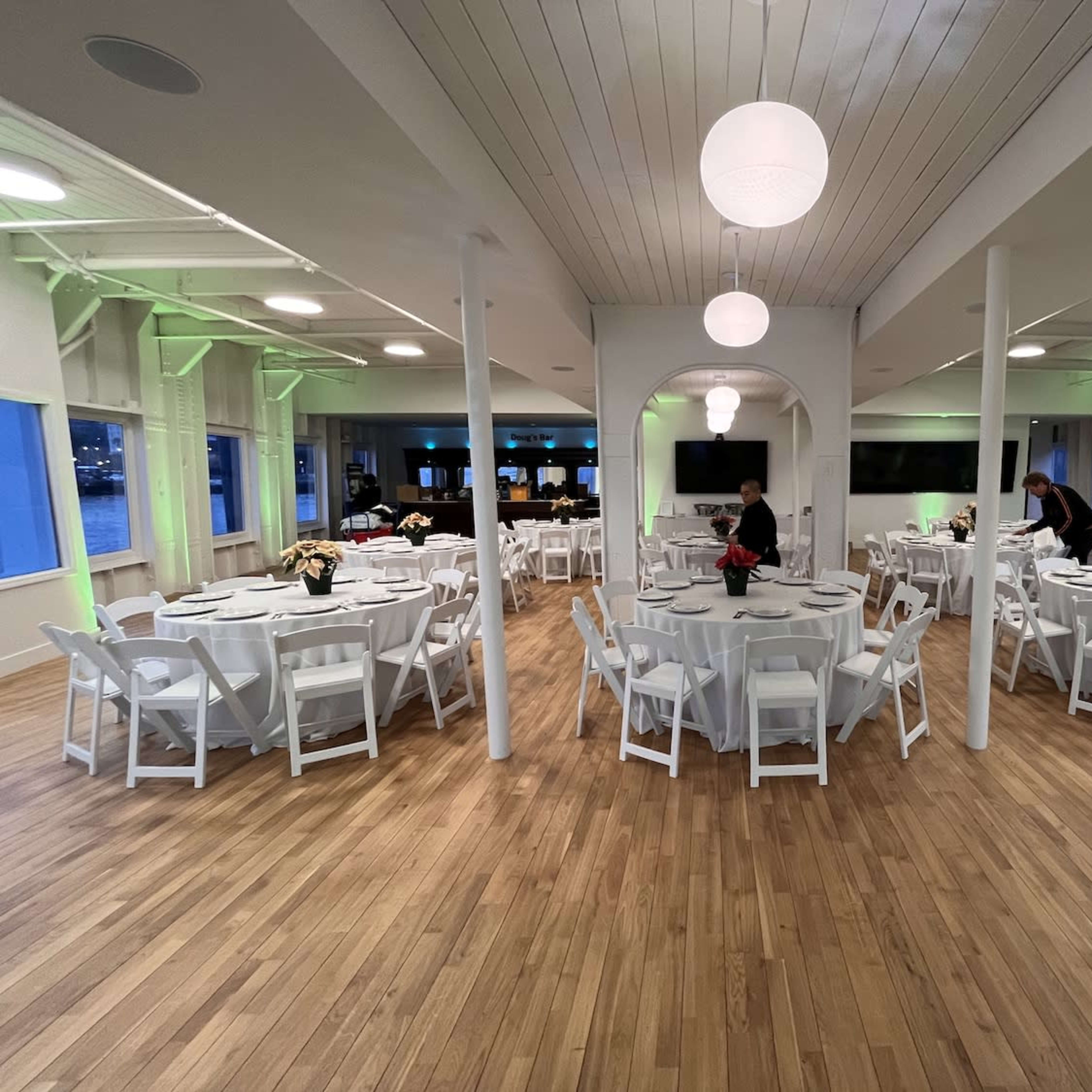 A spacious event venue with round tables set for dining, surrounded by walls with large windows and a polished wood floor.