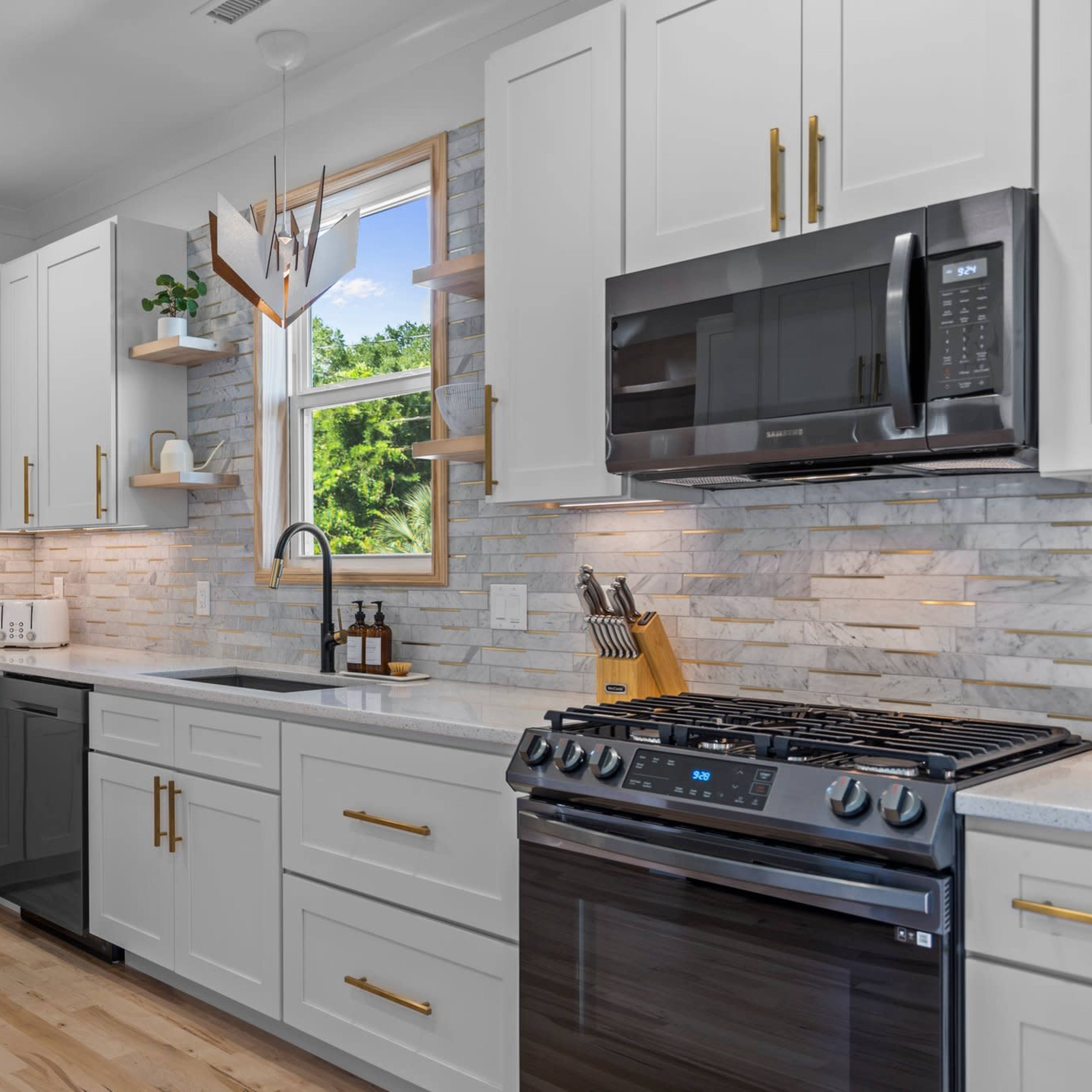 A modern kitchen with white cabinetry, a stainless steel gas range, and a large window above the sink, showcasing stone backsplash and wooden flooring.