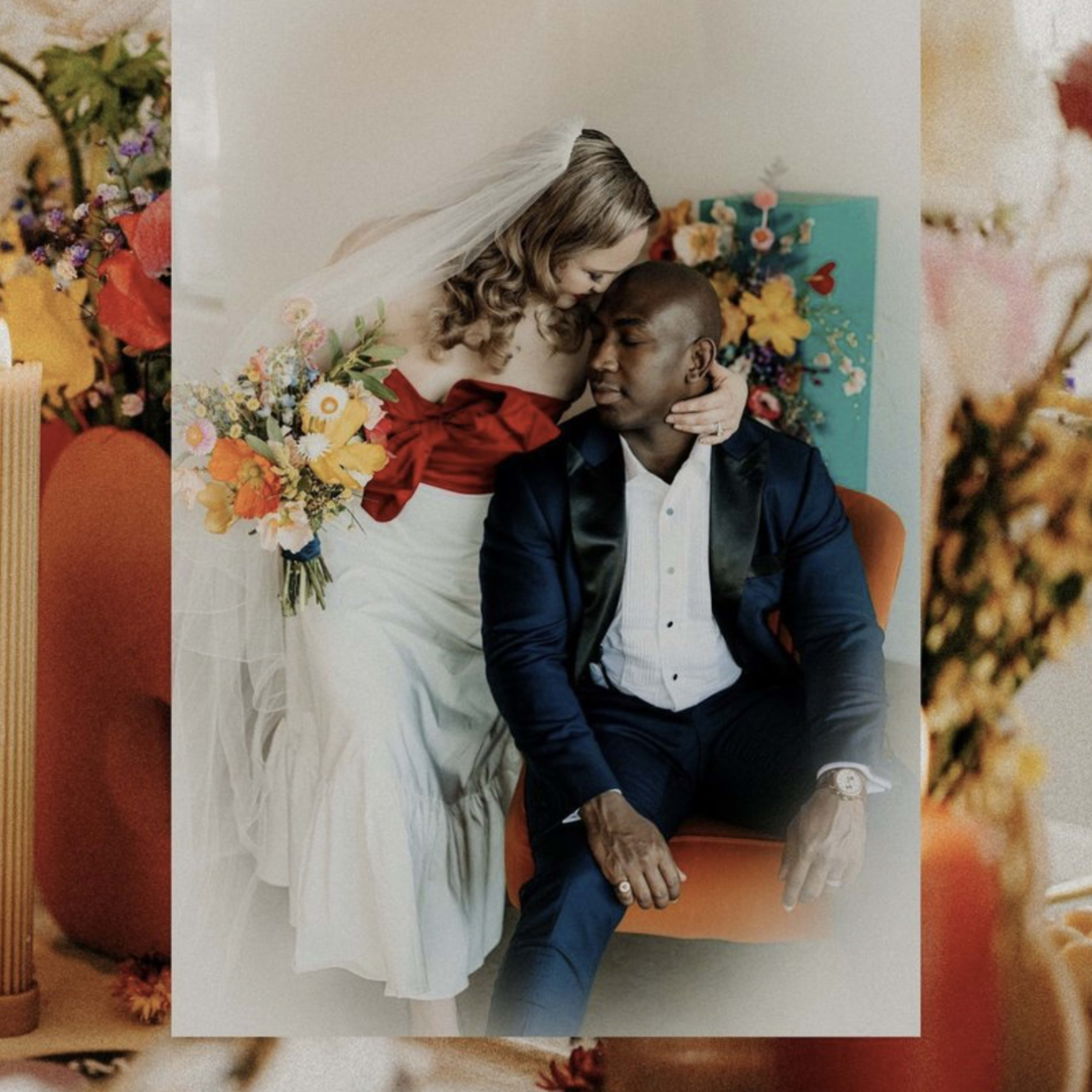 A bride in a white dress with a red sash leans affectionately towards a man in a dark suit while seated against a backdrop of colorful flowers.