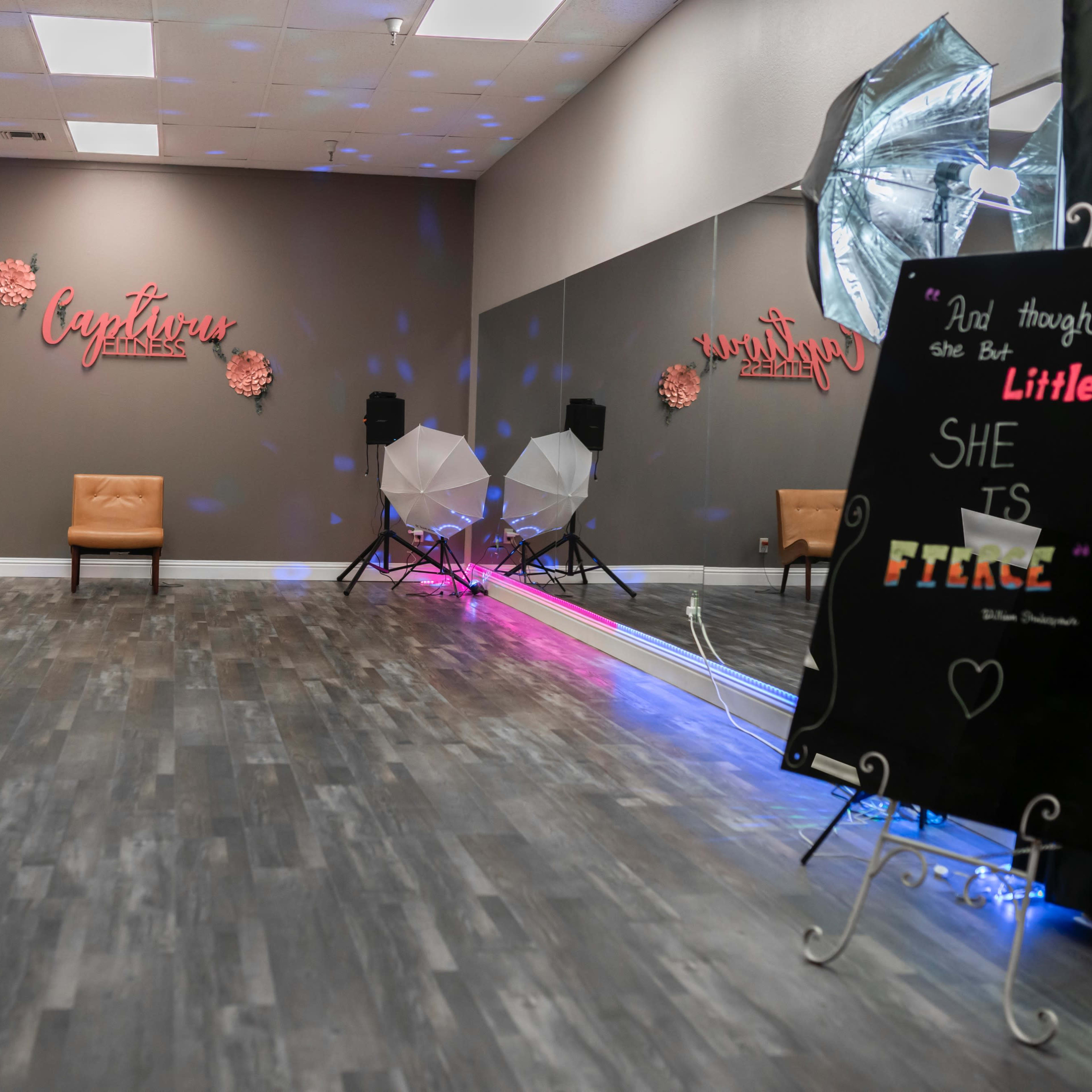 An empty studio space with a brown accent wall, a tan chair, lighting equipment, and a decorative sign that reads, "And though she is little, she is fierce."