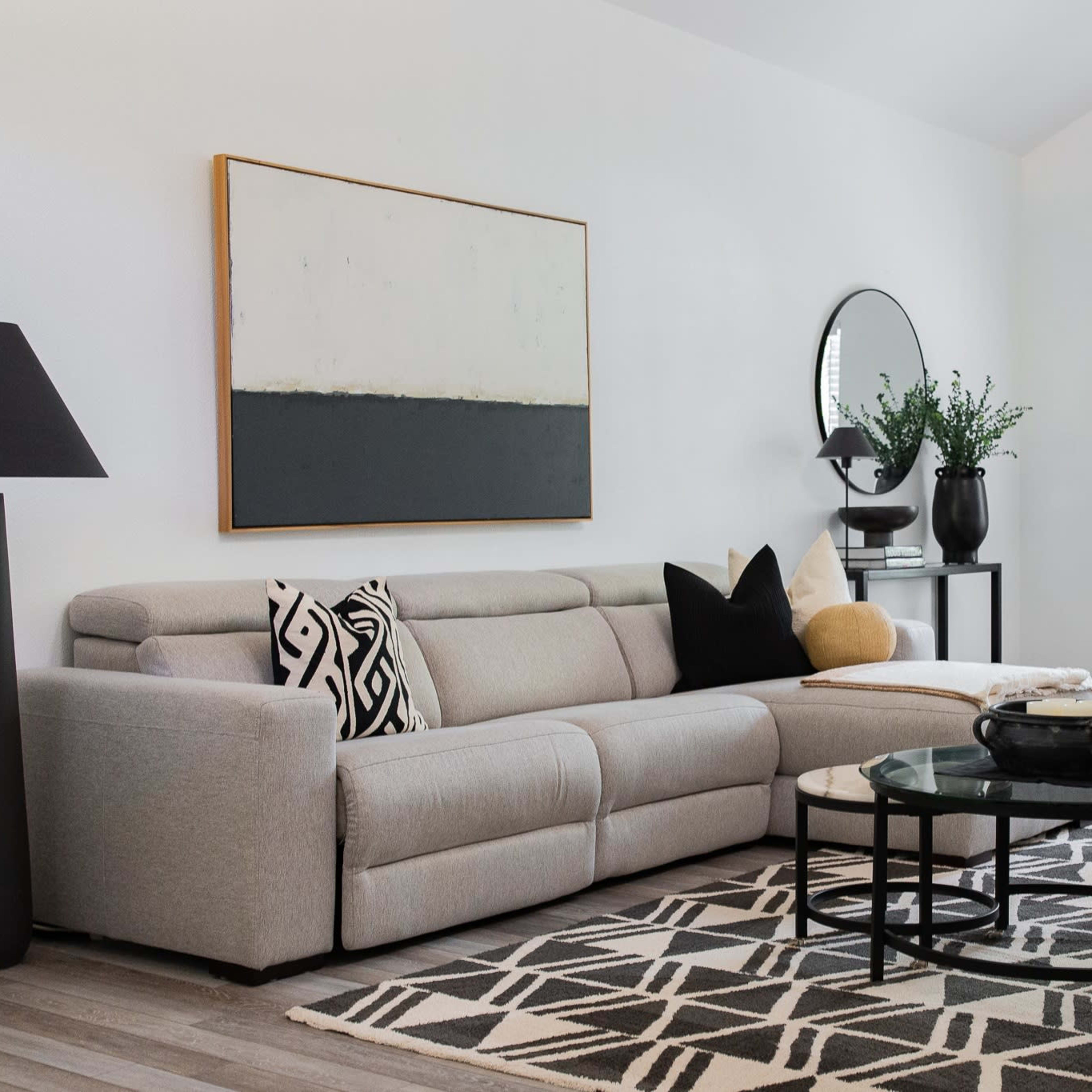 A modern living room features a gray sectional sofa, a black floor lamp, a round glass coffee table, and two white armchairs beside a large window.