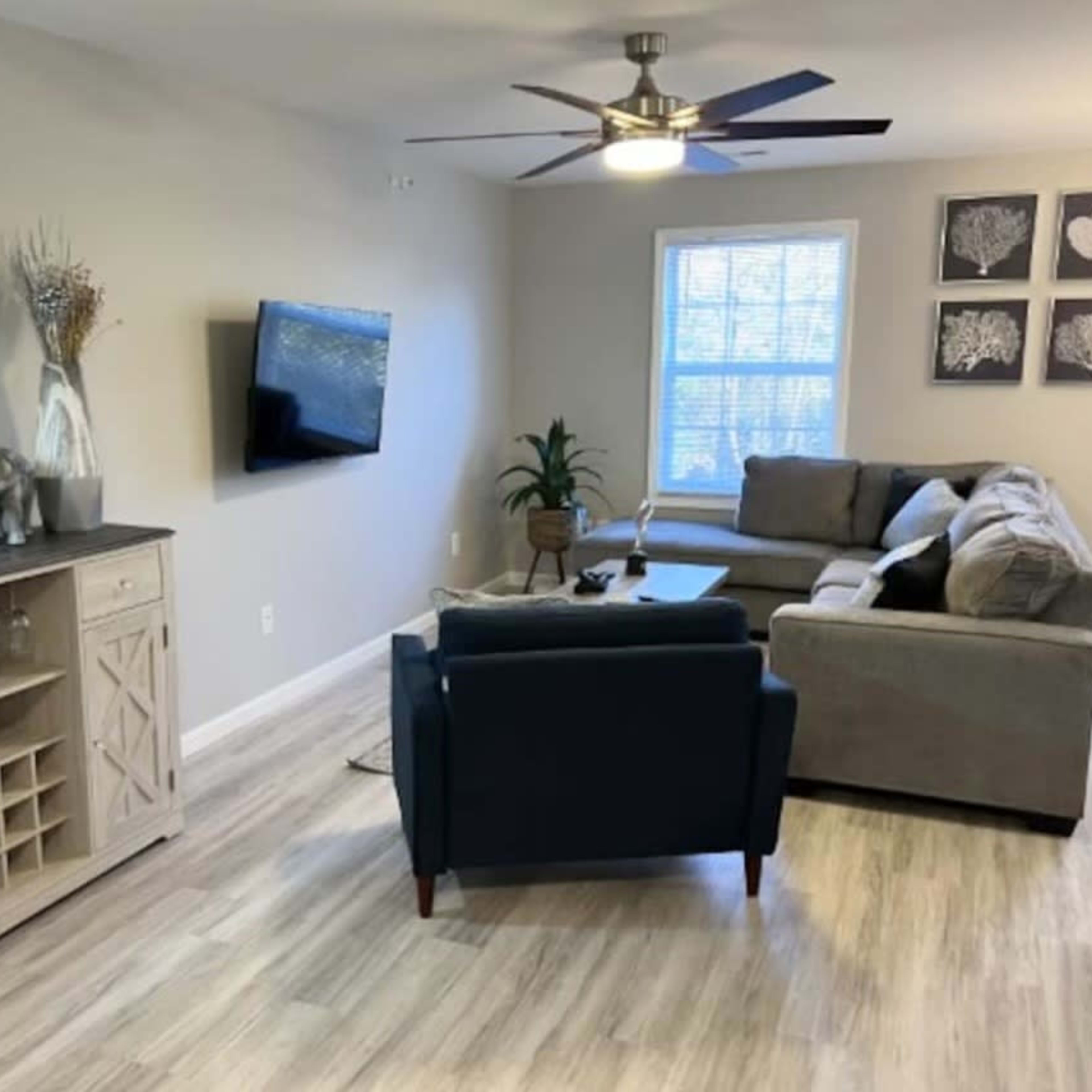 The living room features a comfortable seating arrangement with a gray sectional sofa, a dark blue armchair, a wooden console table, a television mounted on the wall, and decorative art pieces.