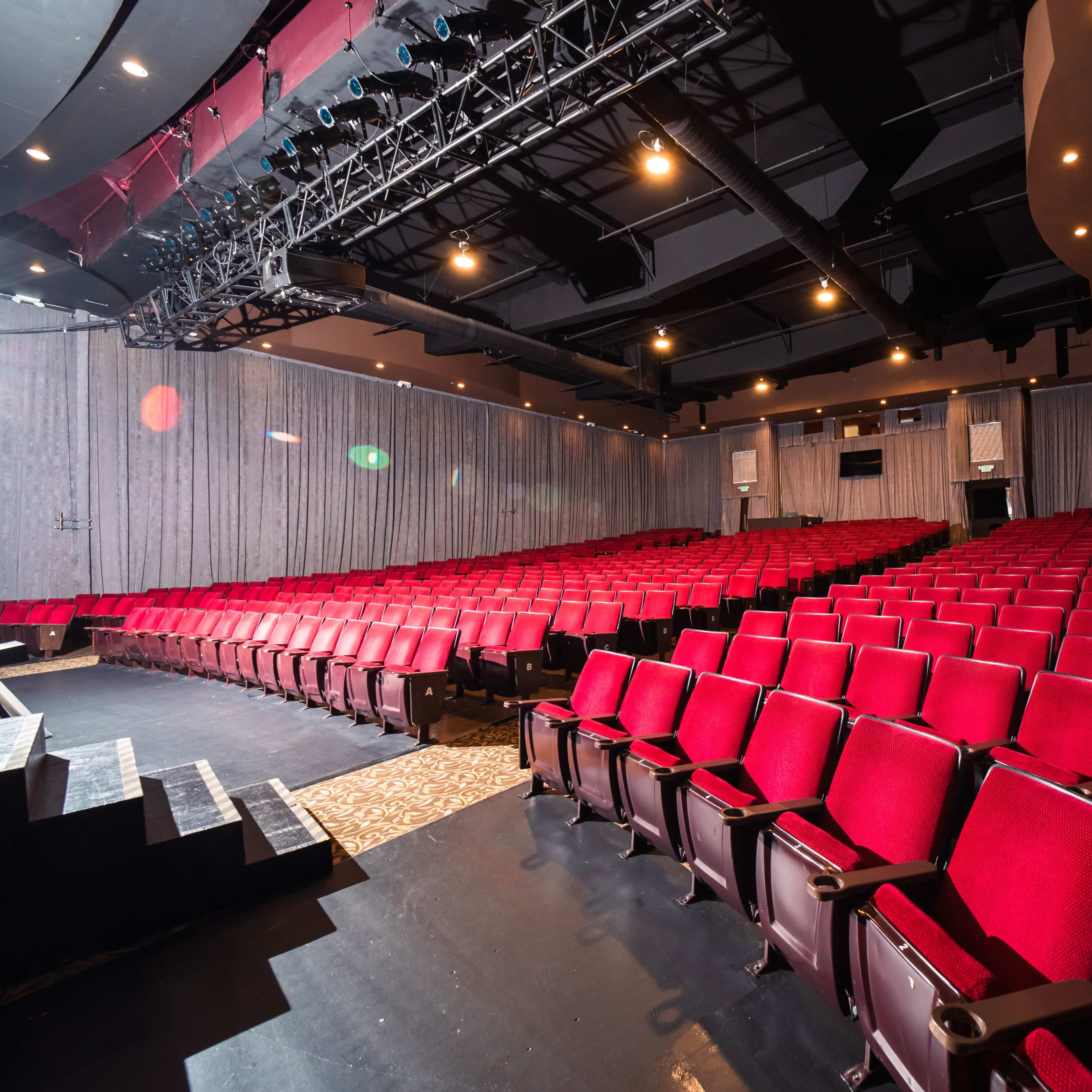 An empty theater with red seats arranged in rows, facing a stage illuminated by overhead lights.