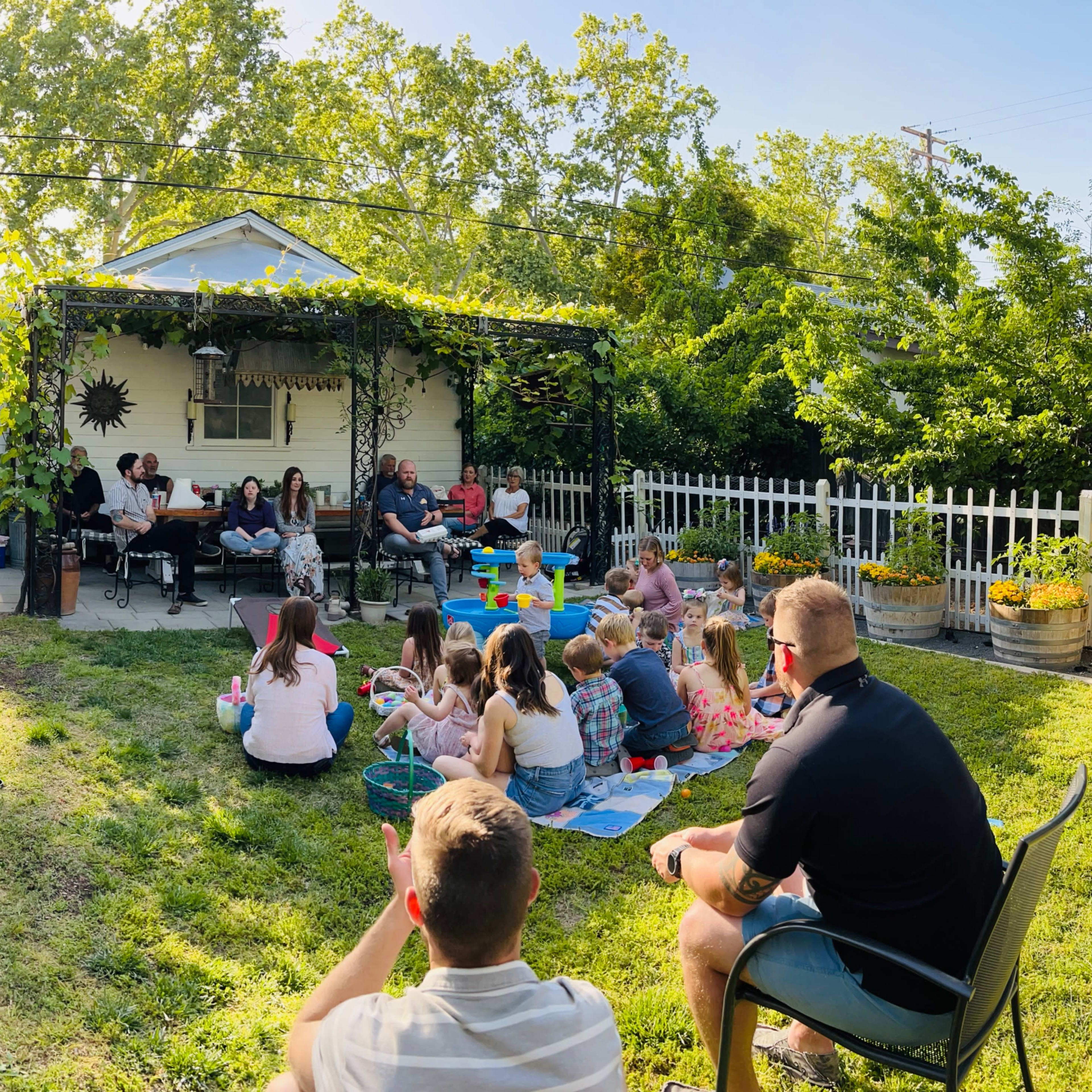 A large gathering of adults and children sits on a grassy yard in front of a house, with some people on chairs and others on blankets, surrounded by trees and a white picket fence.