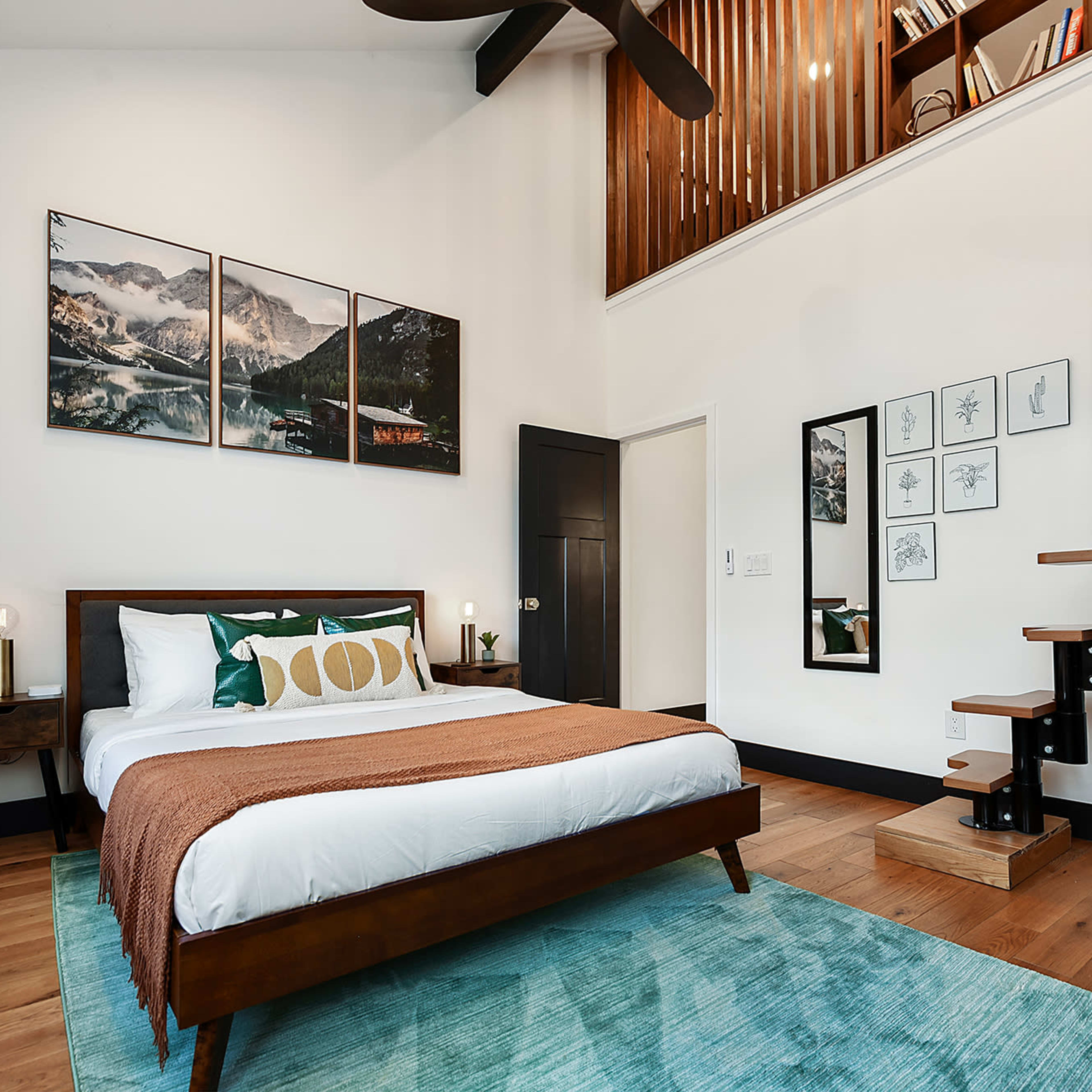 A modern bedroom furnished with a bed, decorative pillows, a bedside table with a lamp, a staircase leading to an upper level, and framed artwork on the walls.