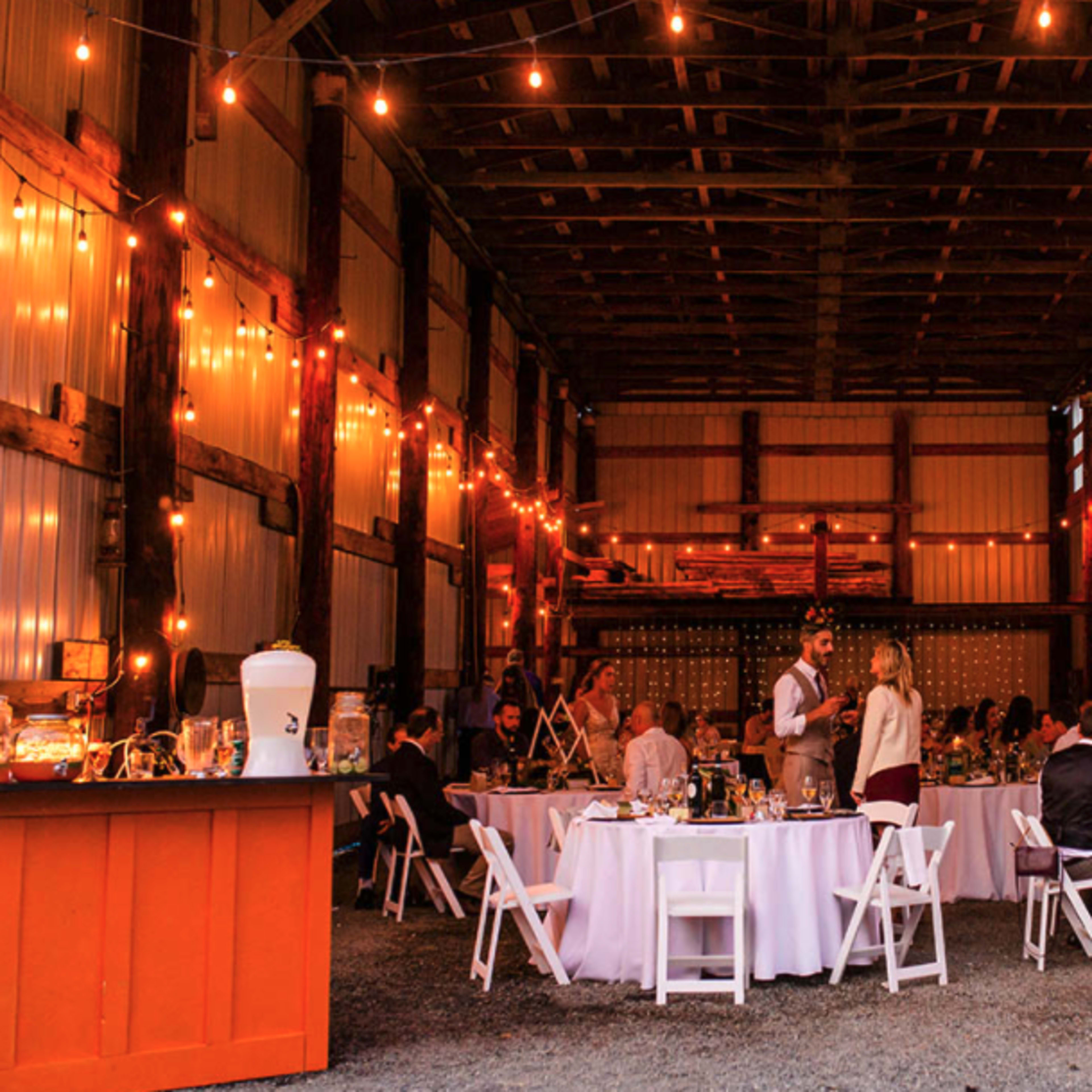 A decorated barn interior with tables set for a wedding reception, string lights illuminating the space, and guests seated at tables.