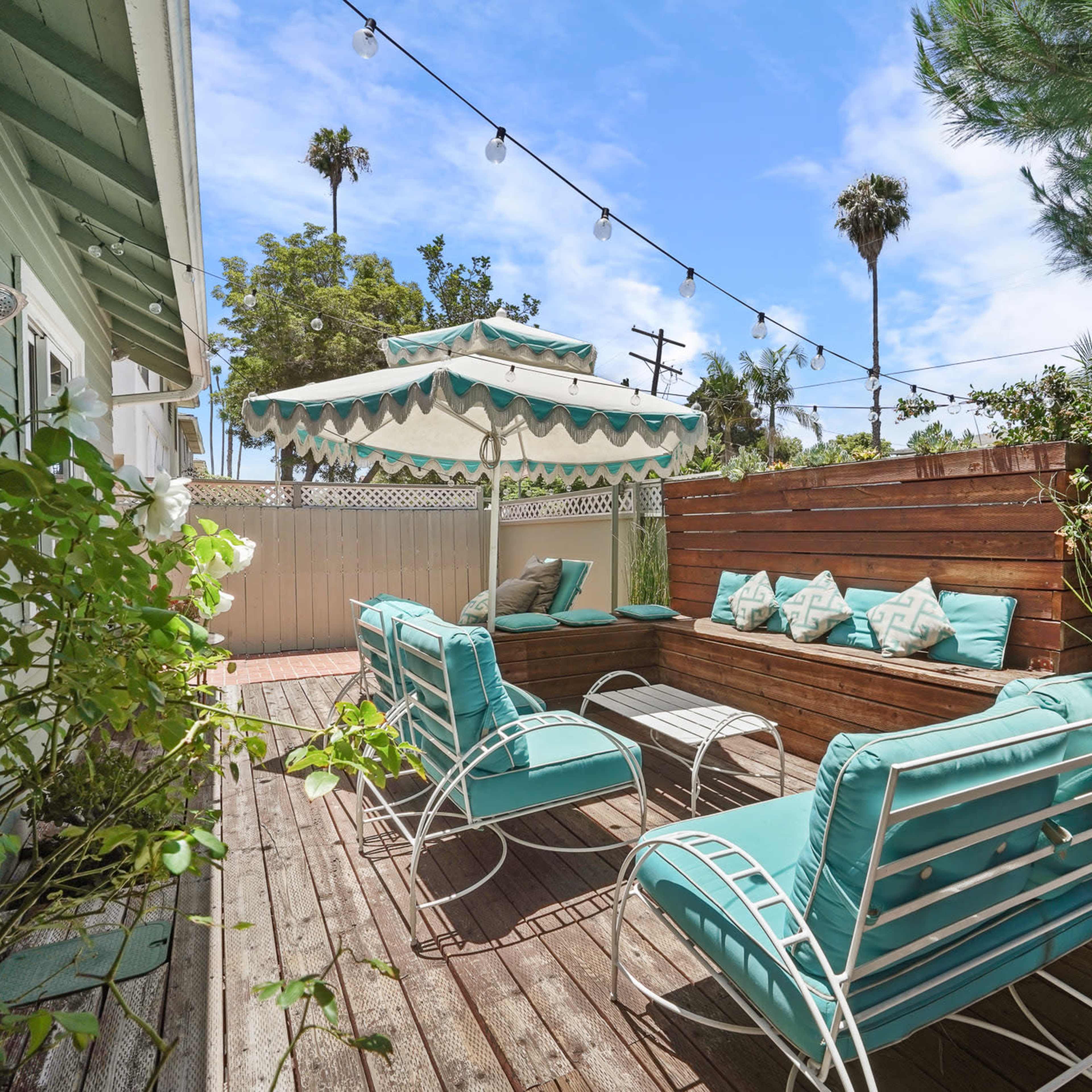 A patio area with turquoise lounge chairs, a table, and a shaded seating area surrounded by greenery and string lights.