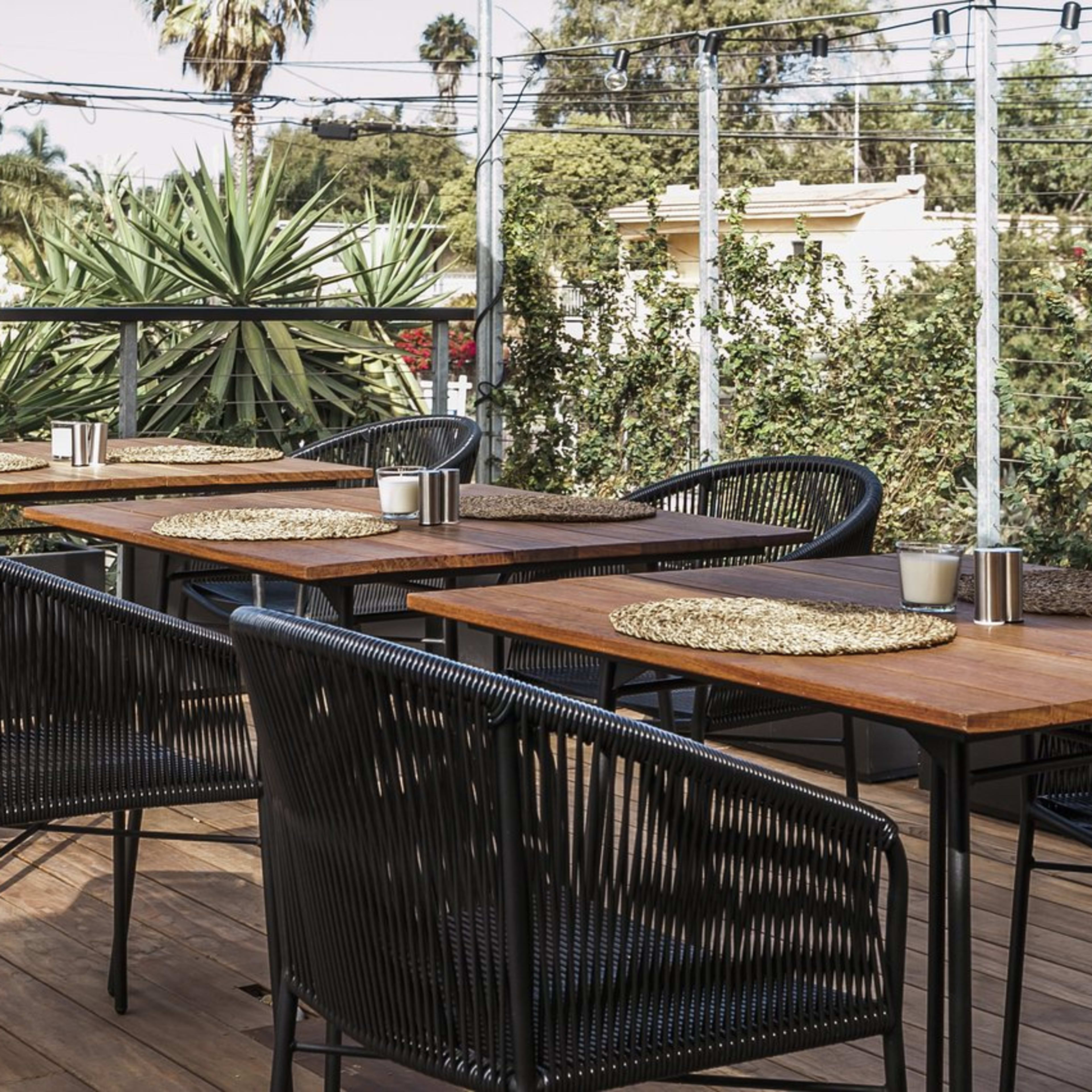 A set of tables and chairs arranged on a wooden deck, surrounded by potted plants and palm trees, with power lines visible in the background.