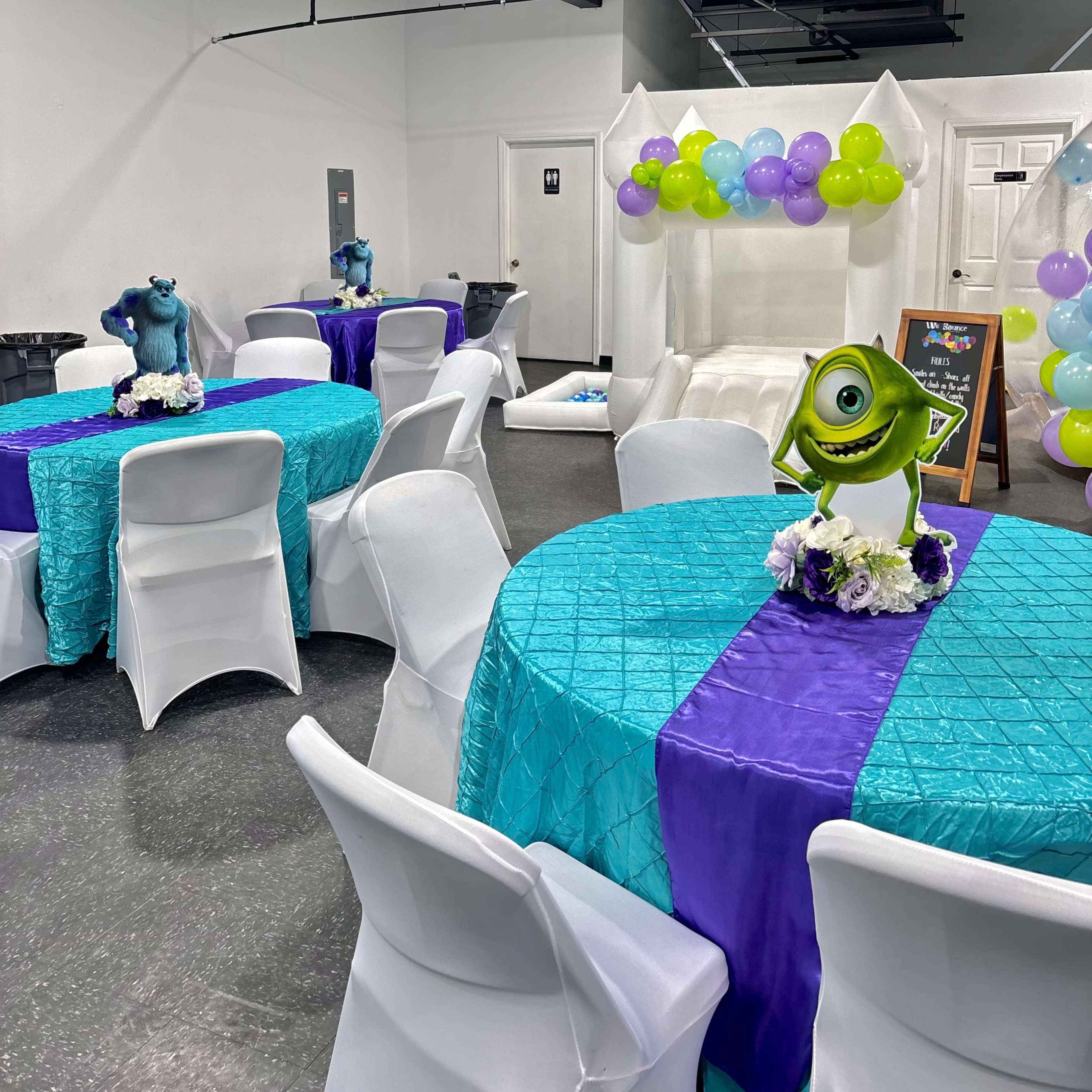 A brightly decorated party room with tables covered in turquoise and purple fabrics, featuring centerpieces of animated characters, and balloons decorating the background.