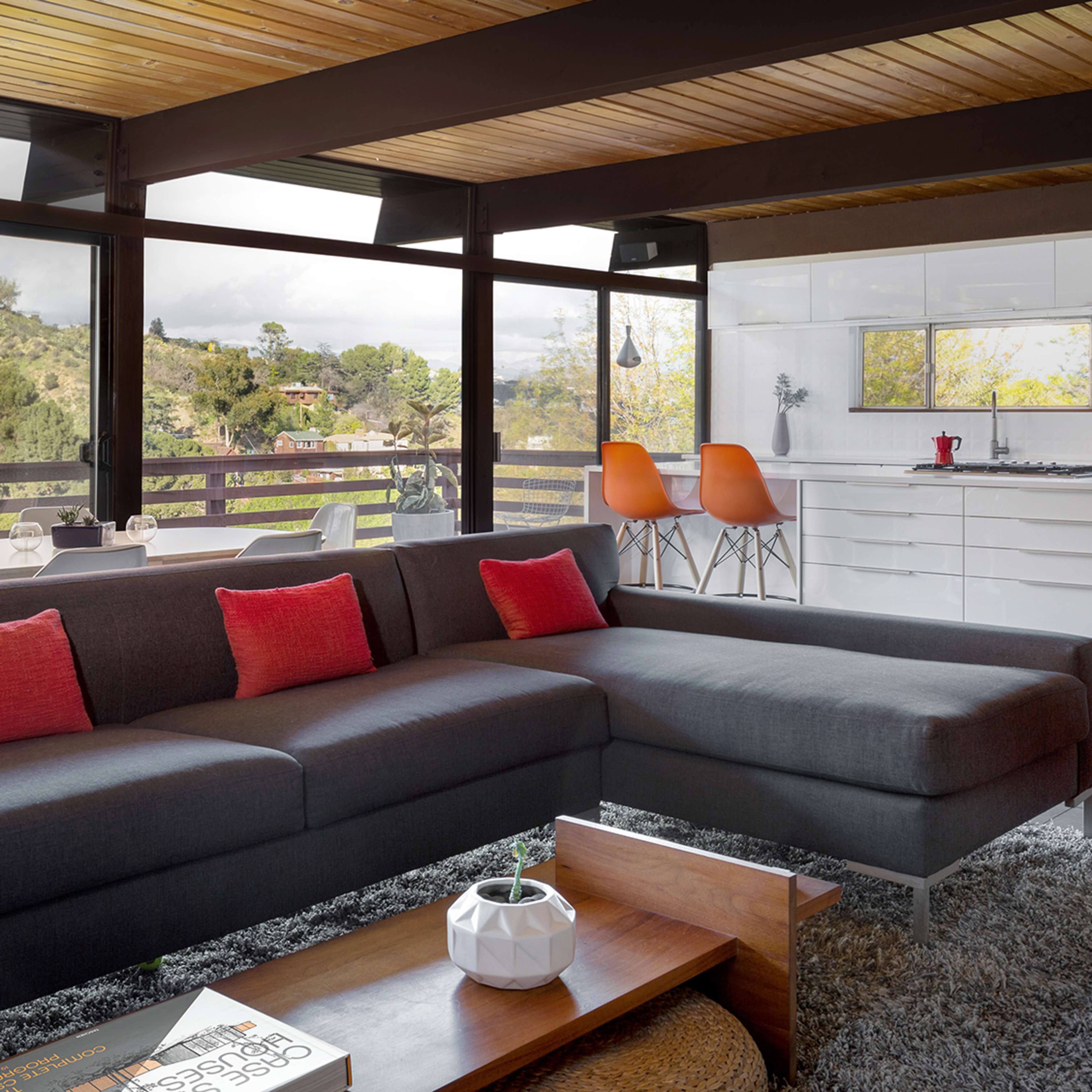 A modern living room features a sectional sofa with red cushions, a wooden coffee table, and large windows that overlook a green hillside and a dining area with a white kitchen.