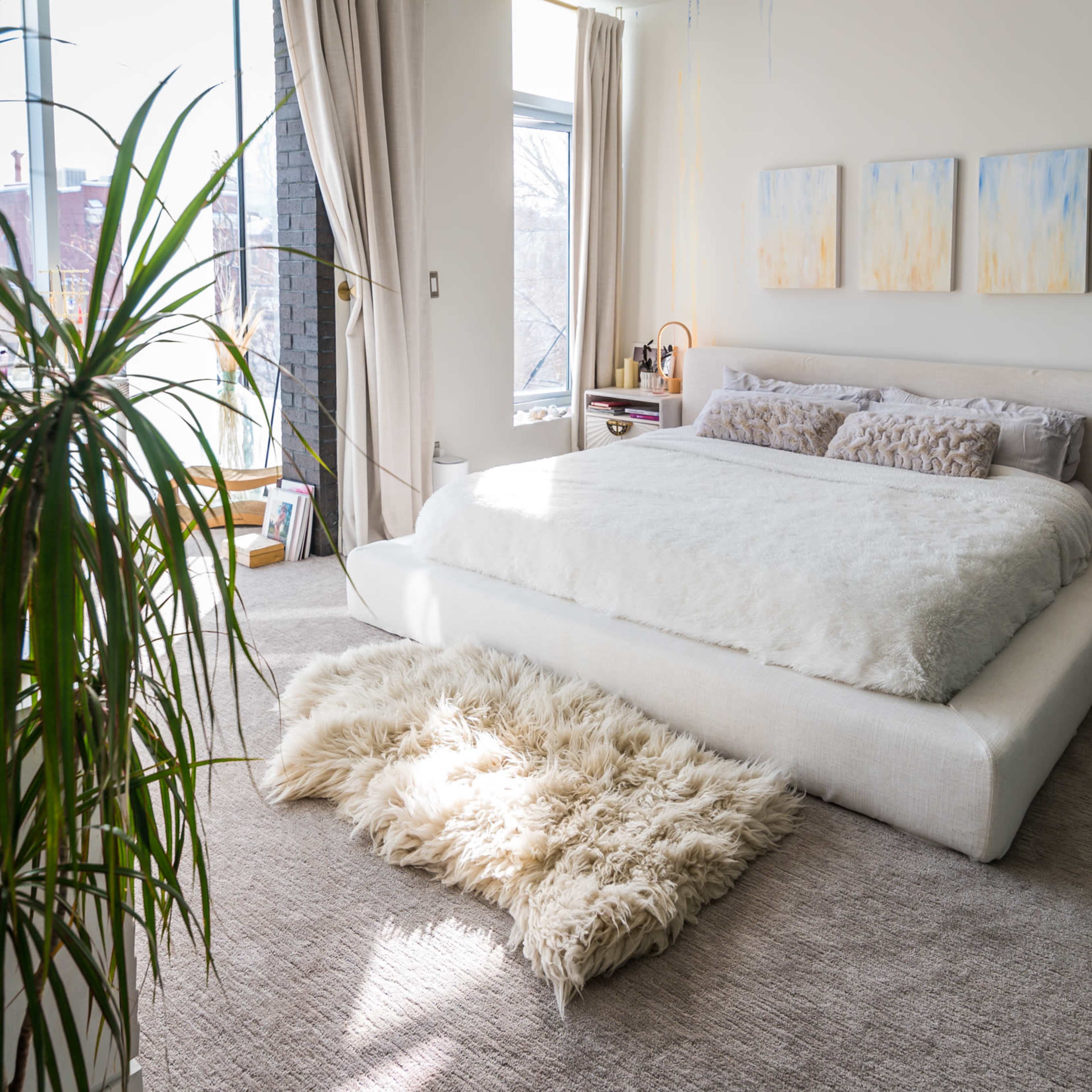 A bedroom featuring a large bed with a white bedspread and decorative pillows, flanked by nightstands and a plush rug on a carpeted floor, illuminated by natural light from large windows.