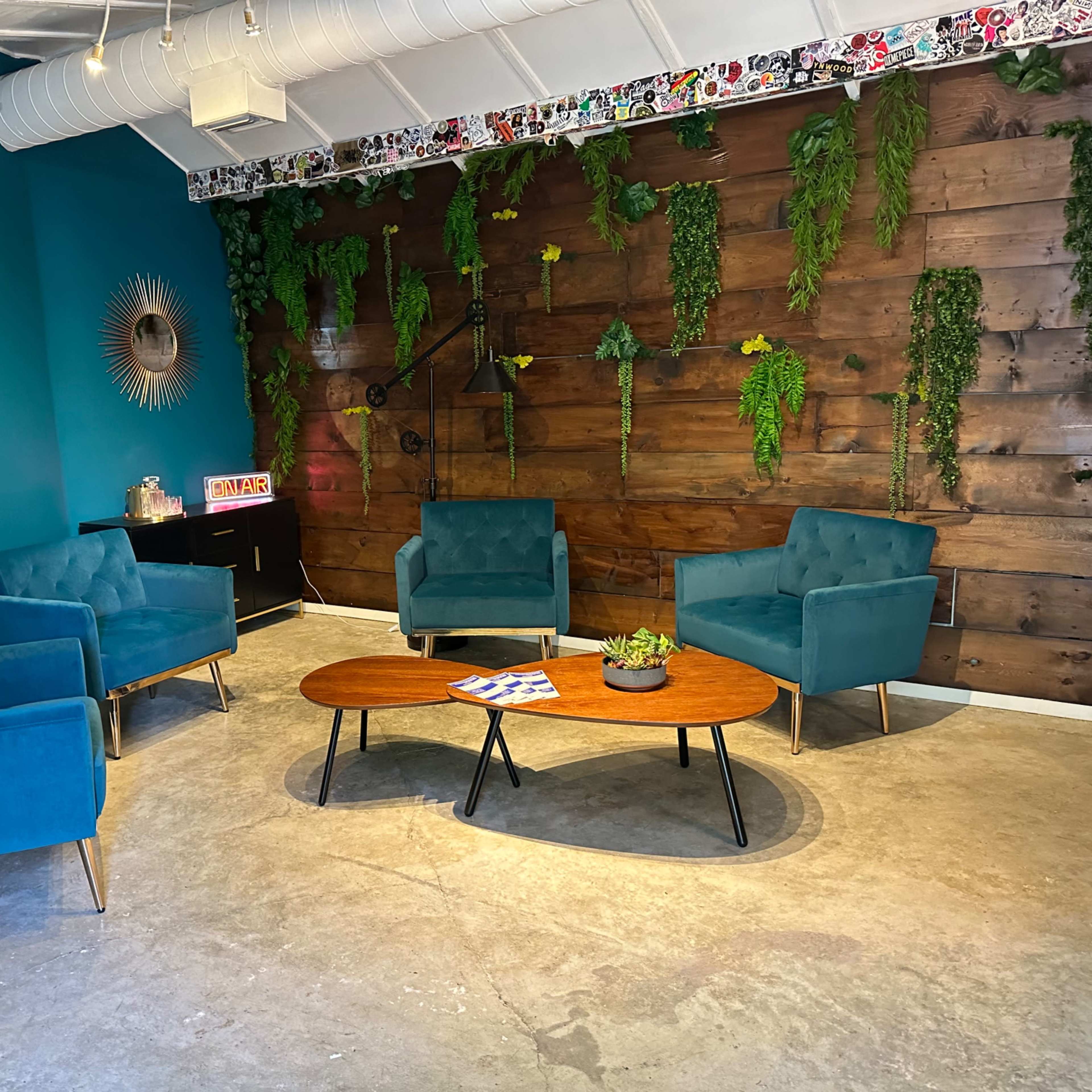 A modern seating area with four teal chairs, a wooden coffee table, and a wall featuring greenery and decorative art in a vibrant blue and brown setting.