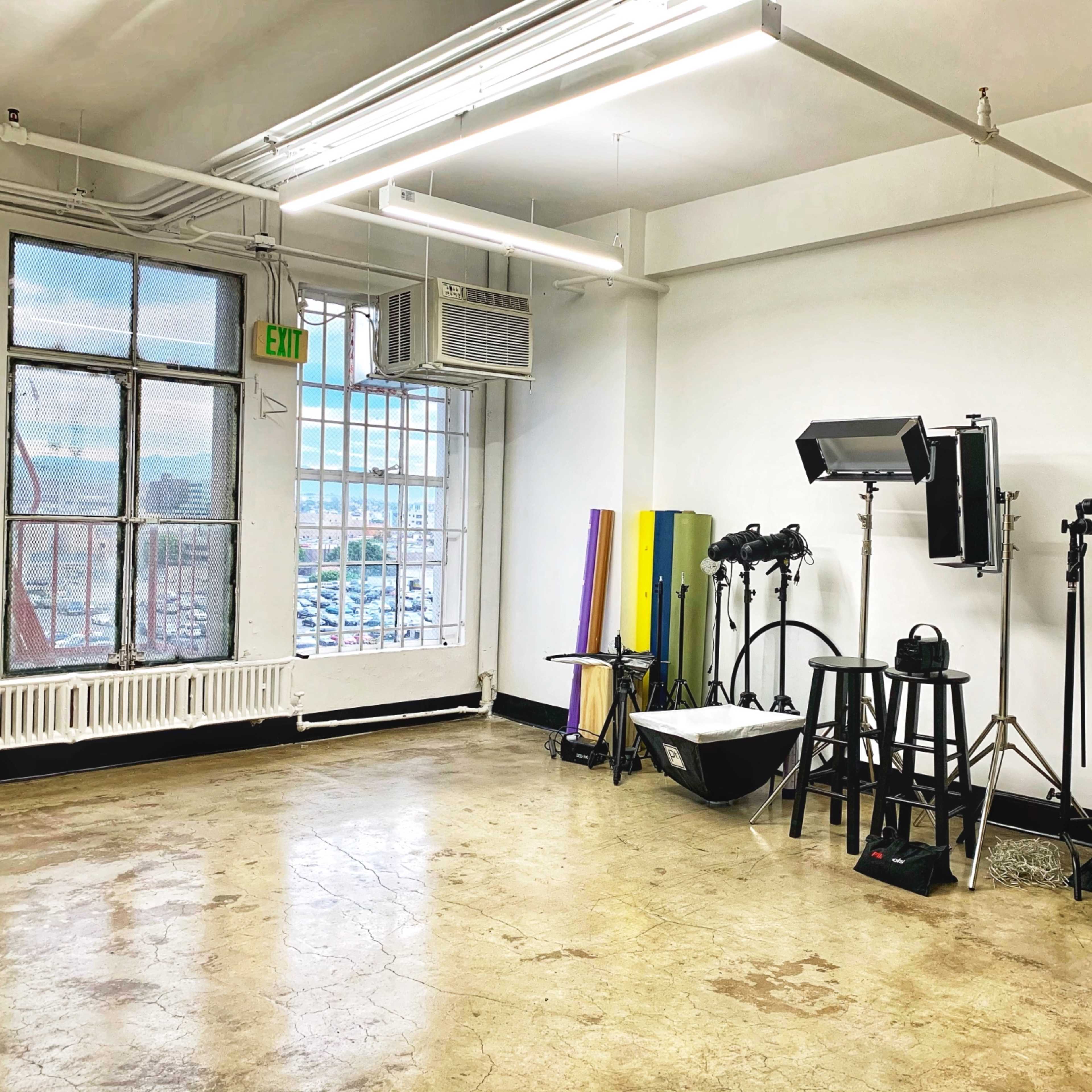 A spacious, brightly lit studio with large windows, a photography setup including lights and stands, and a bare concrete floor.
