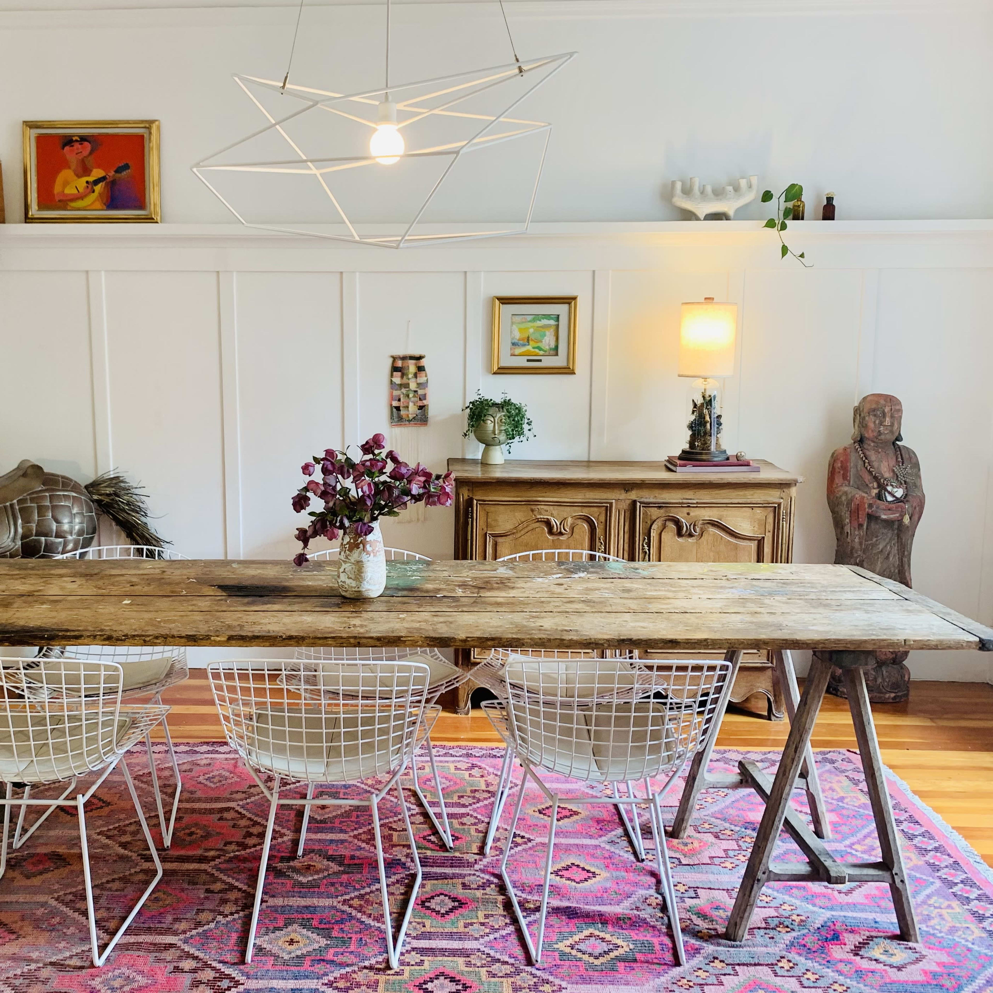 A rustic wooden dining table is surrounded by four white wire chairs in a brightly lit room featuring decorative art pieces, a statue, and an antique sideboard.