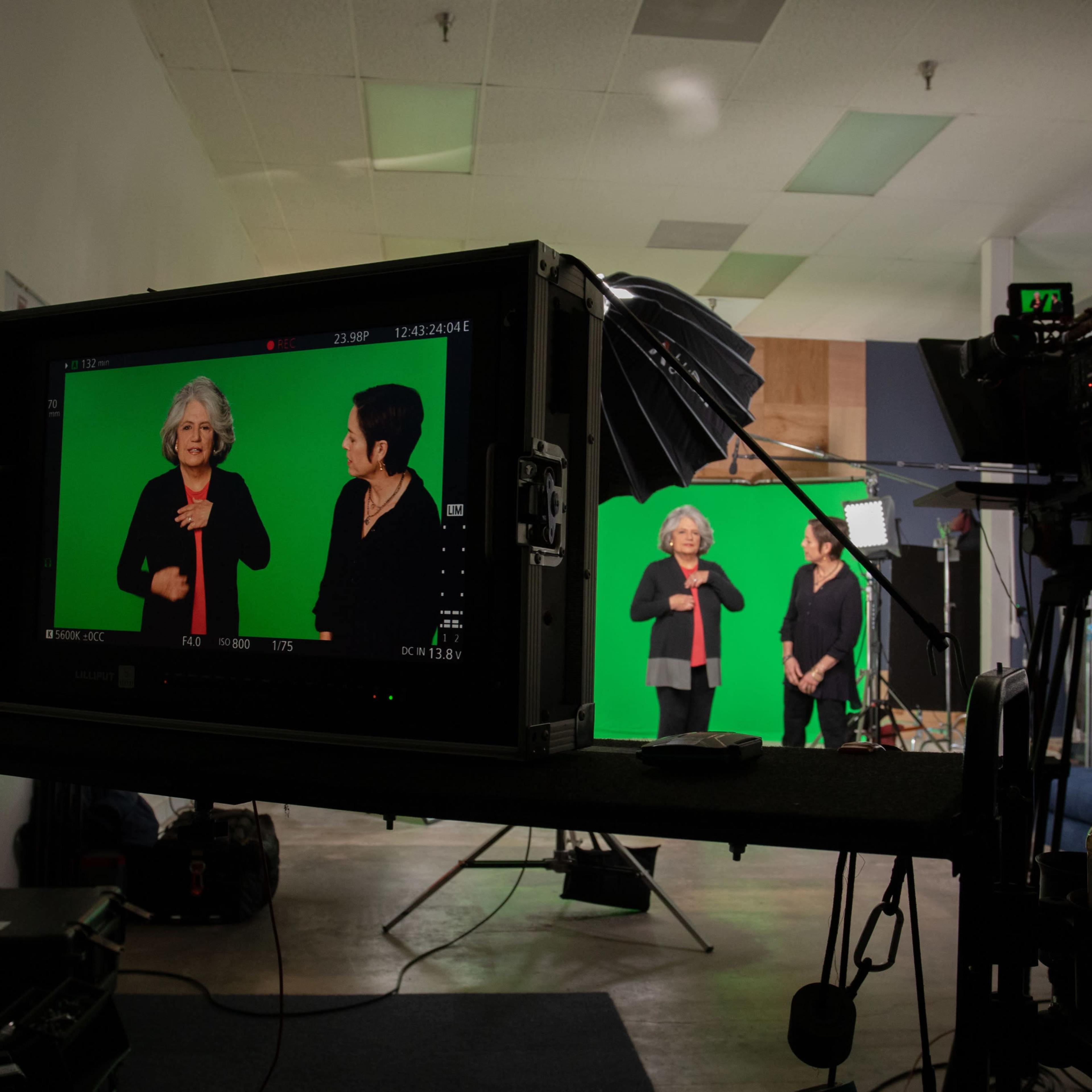 A video recording setup features two women discussing in front of a green screen, with a camera and monitor in the foreground.