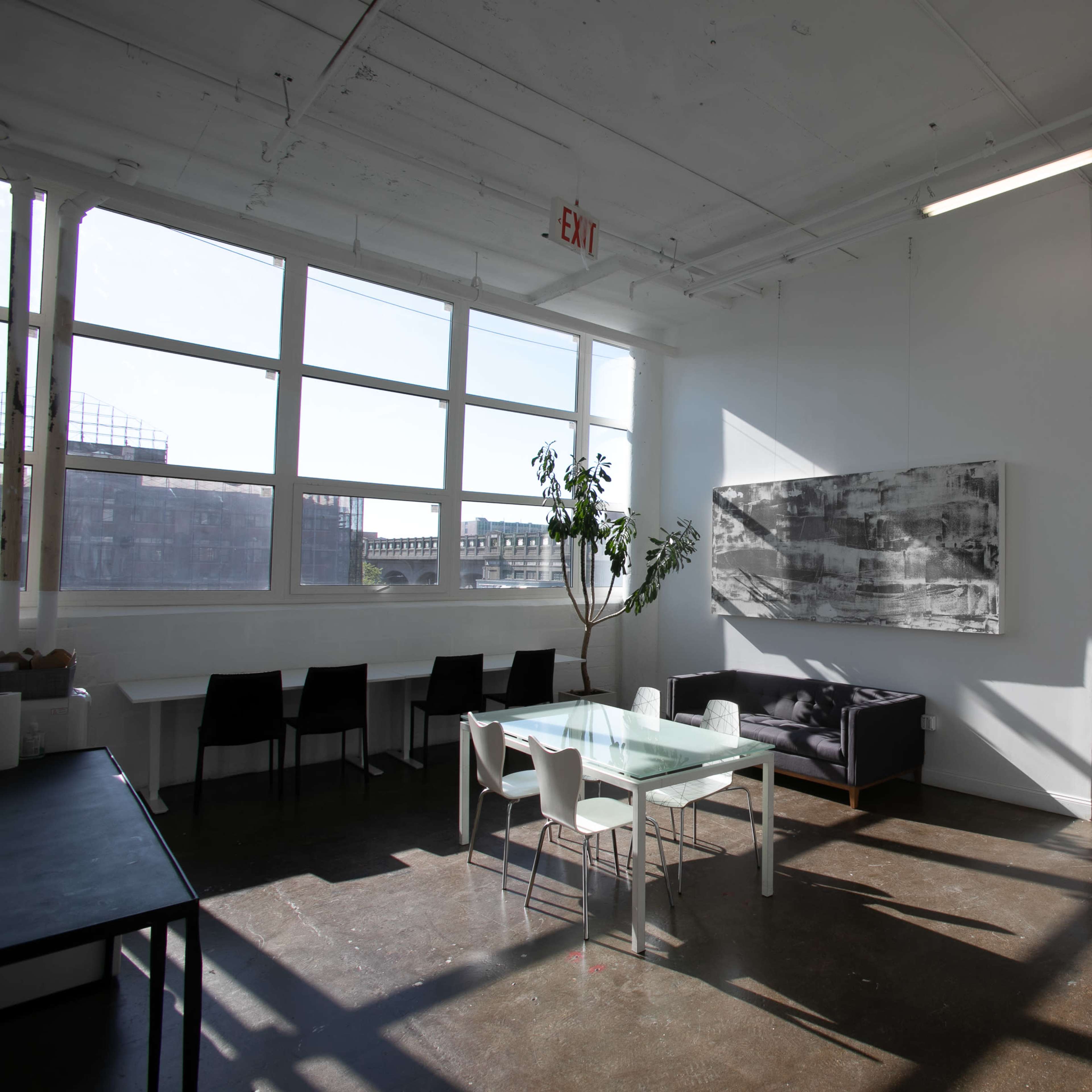A modern, open office space features large windows, a dining table, chairs, and artwork on the wall.
