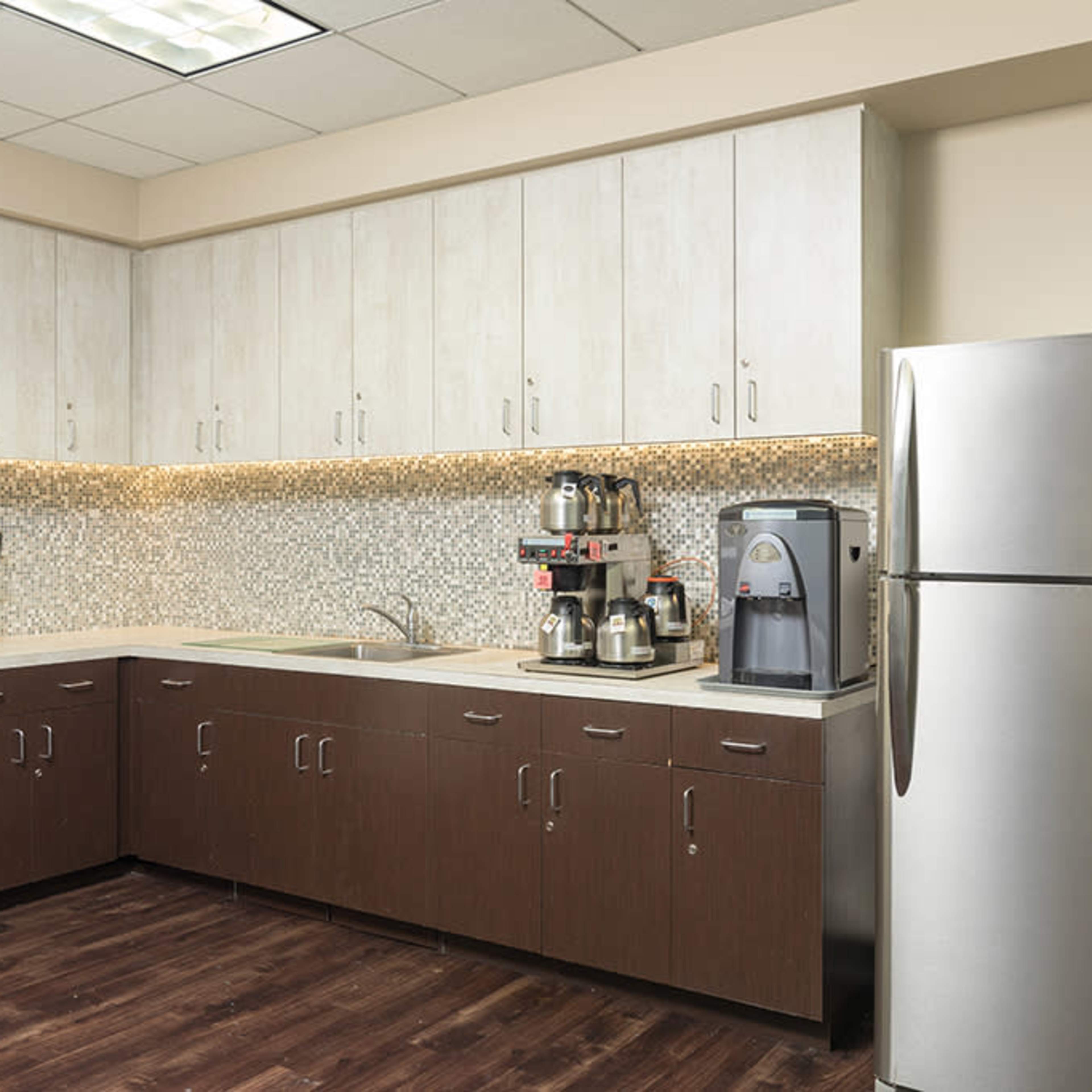 A modern kitchen area features glossy white cabinets, a silver refrigerator, a coffee station, and various appliances against a backsplash of mosaic tiles.