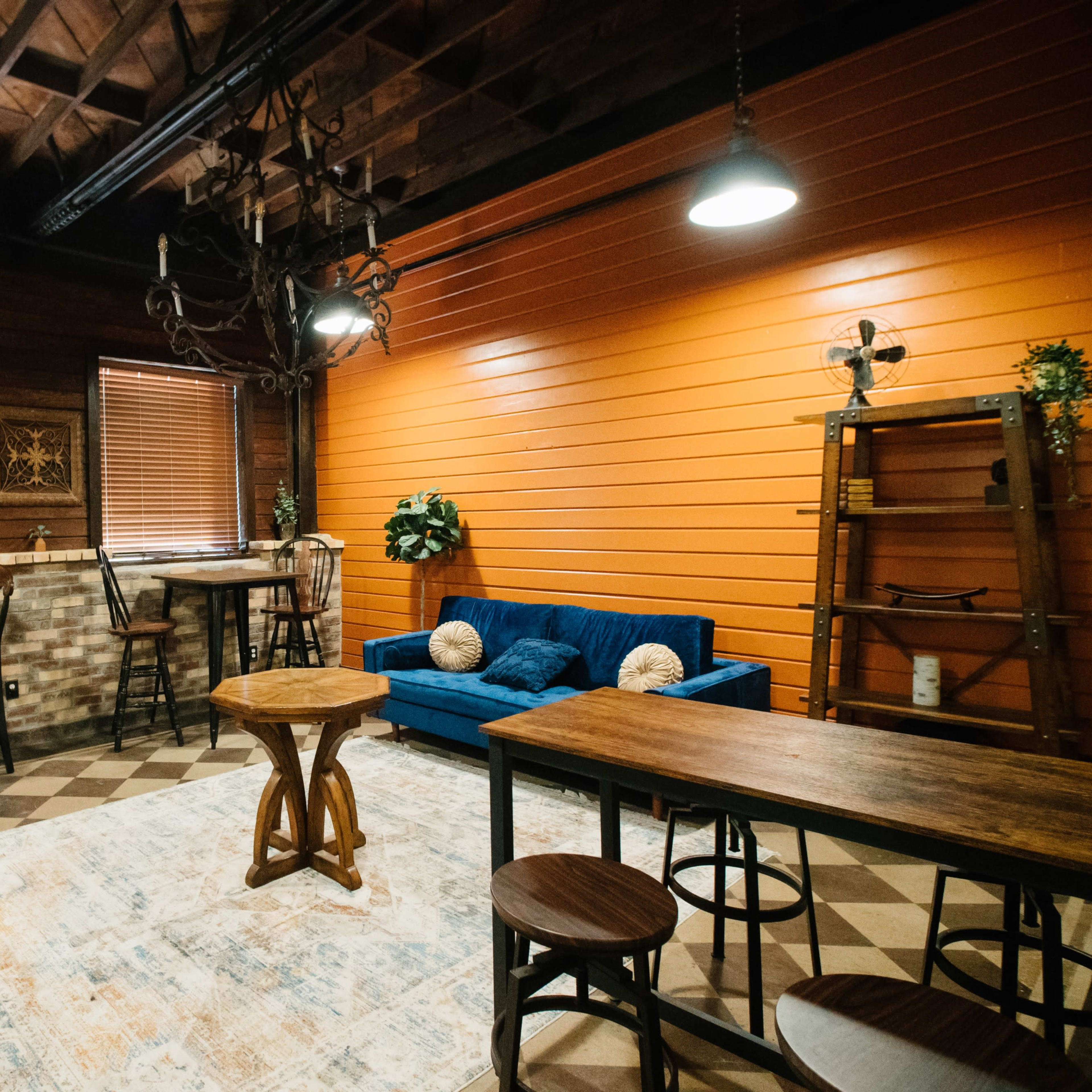 A cozy room featuring a blue couch, several wooden tables and chairs, a patterned rug, and orange accent walls with window blinds.