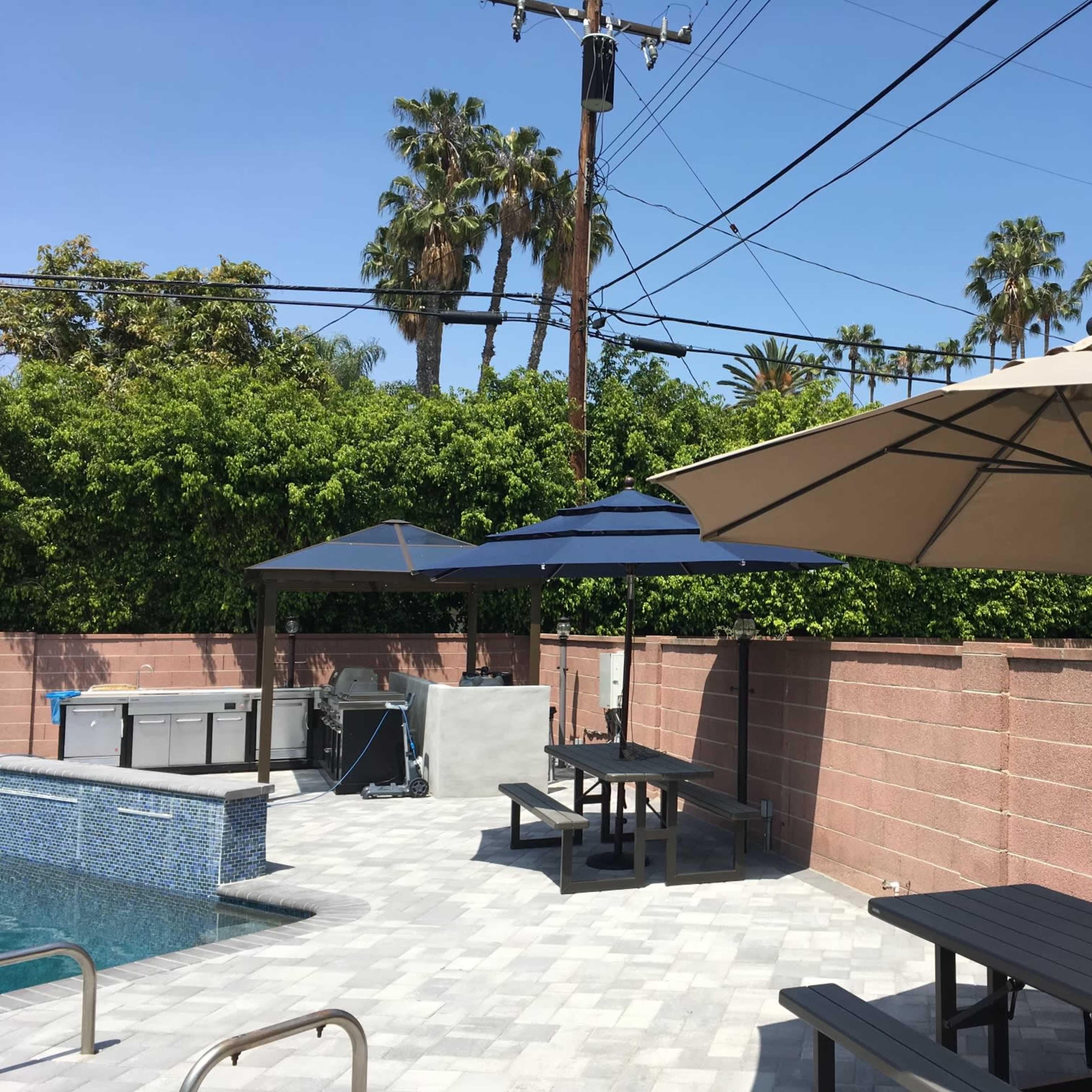 A patio area with two large umbrellas, a rectangular pool, several tables, and palm trees in the background under a clear blue sky.