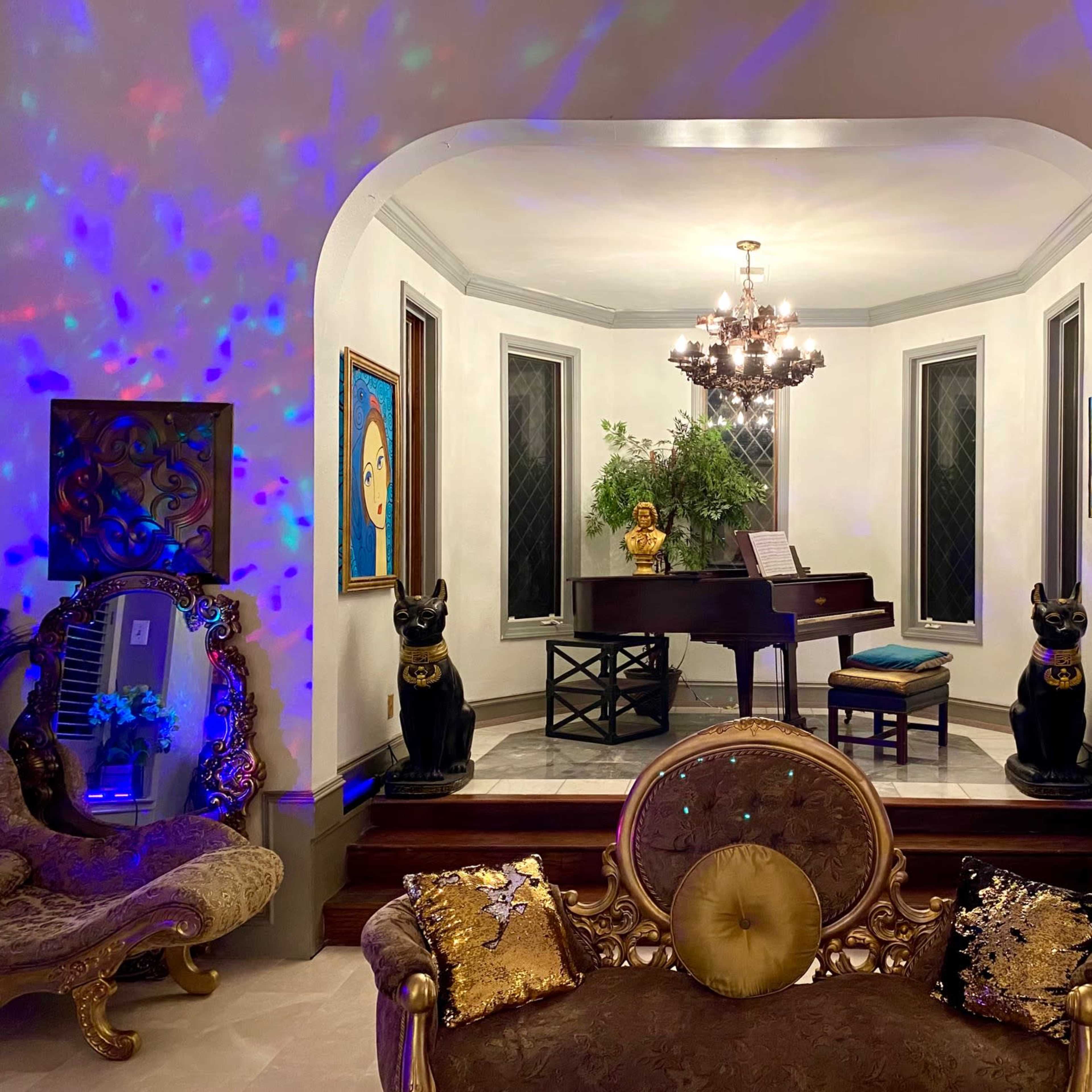 A grand room features a piano at the center, flanked by decorative statues and illuminated by a chandelier, with colorful lights reflecting off the walls.