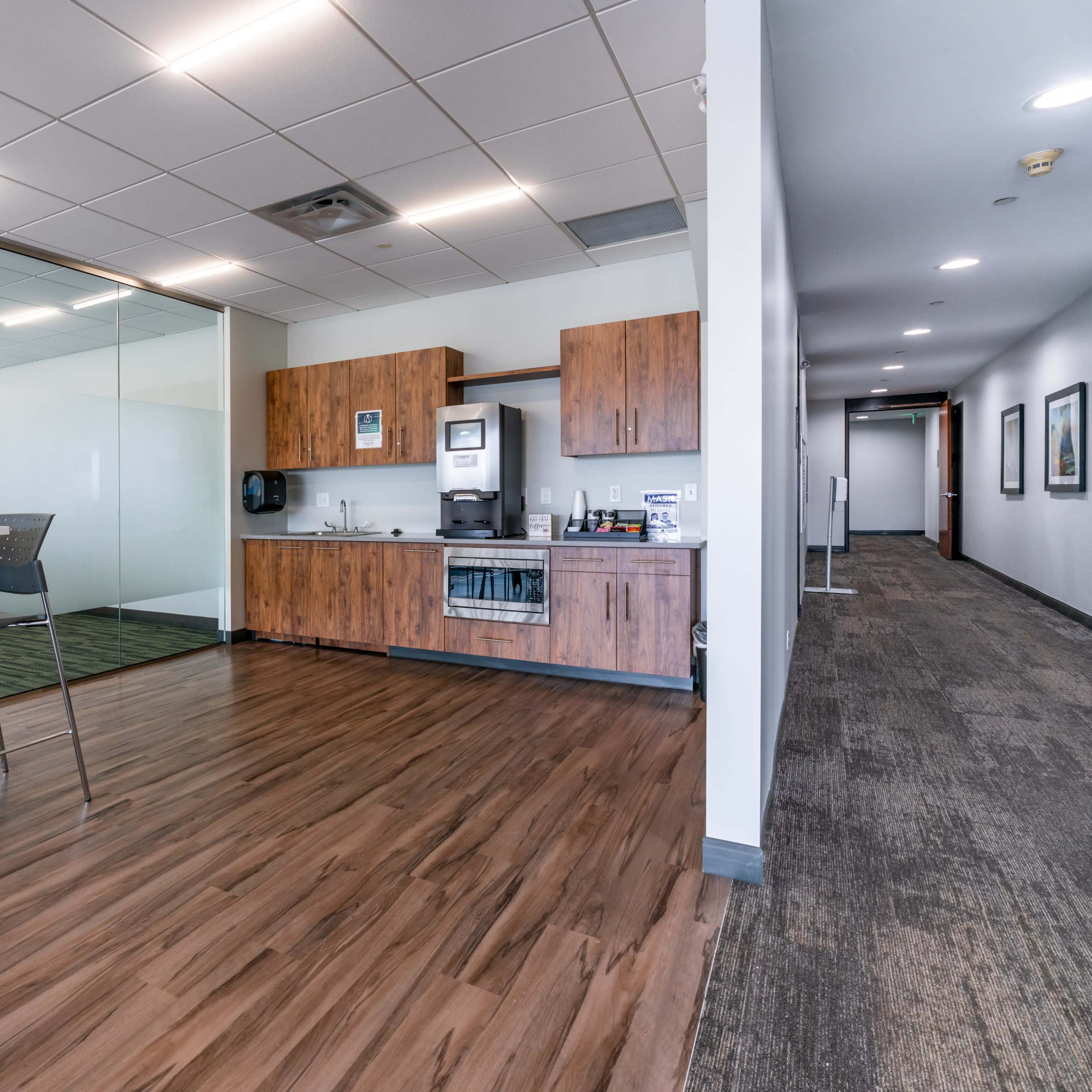 A modern office break area with a kitchenette, tall table and chairs, and a hallway leading to closed office spaces.