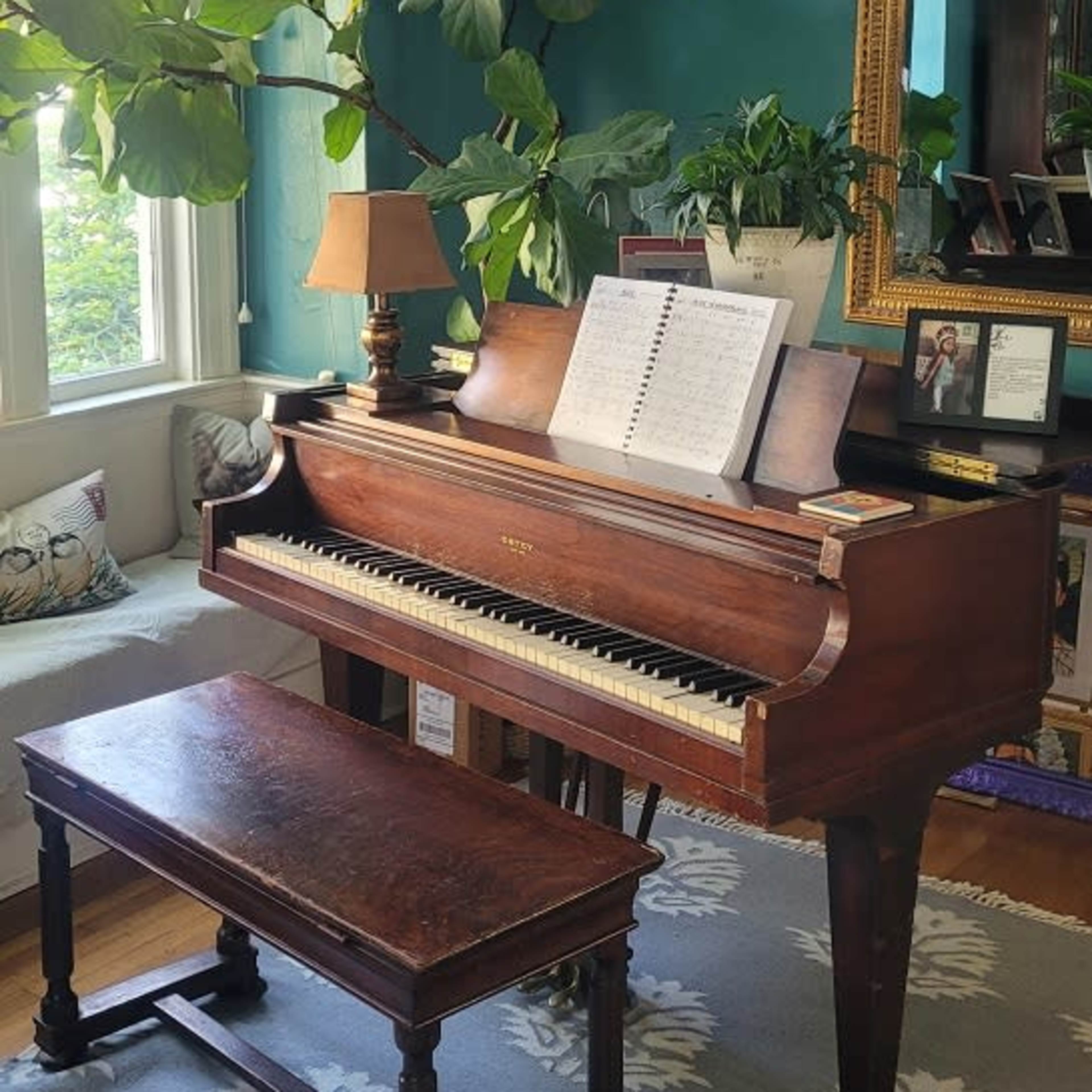 A wooden piano with sheet music sits next to a bench in a room with green walls and large windows, surrounded by plants and a mirror.