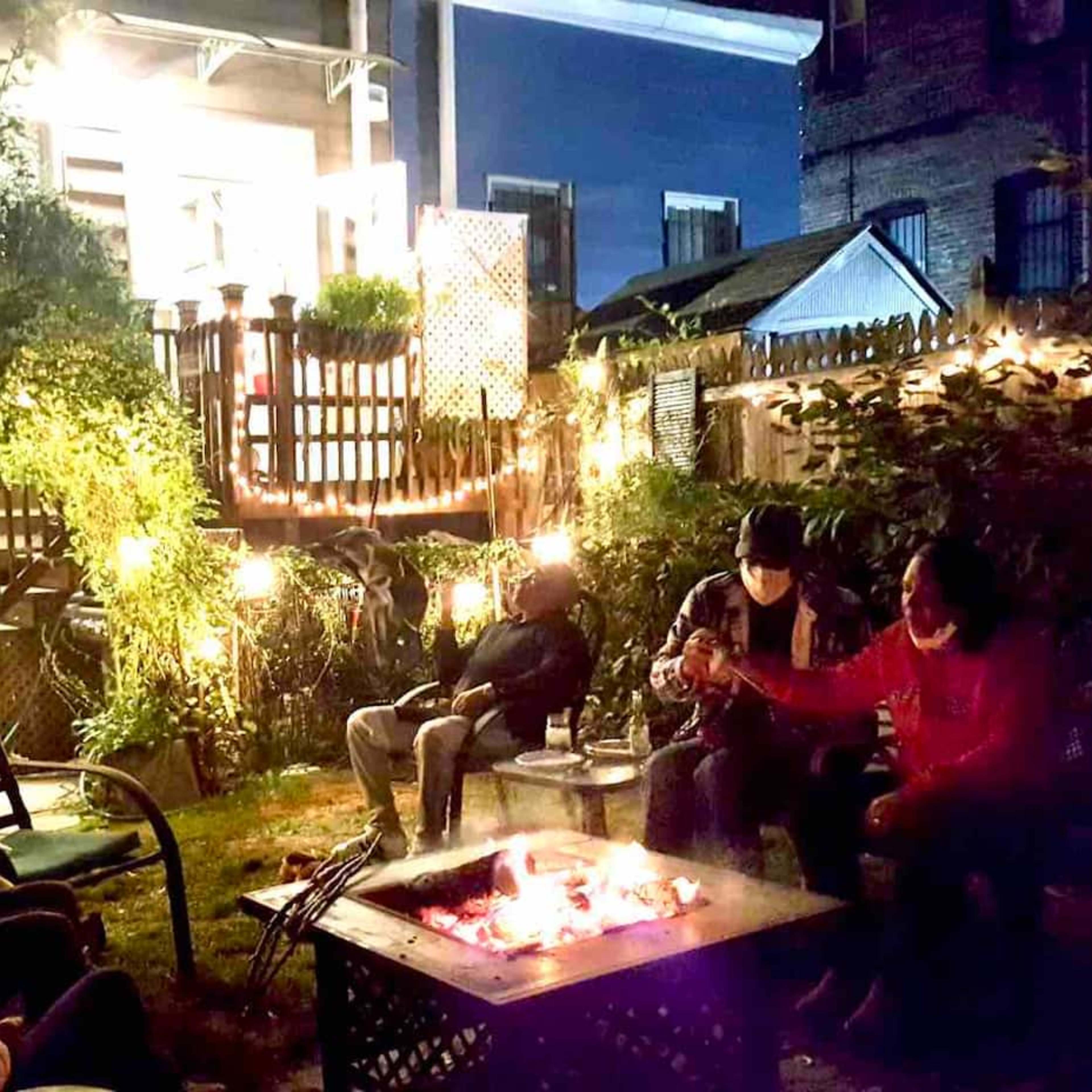 A group of people sits around a fire pit in a well-lit backyard, surrounded by greenery and string lights.
