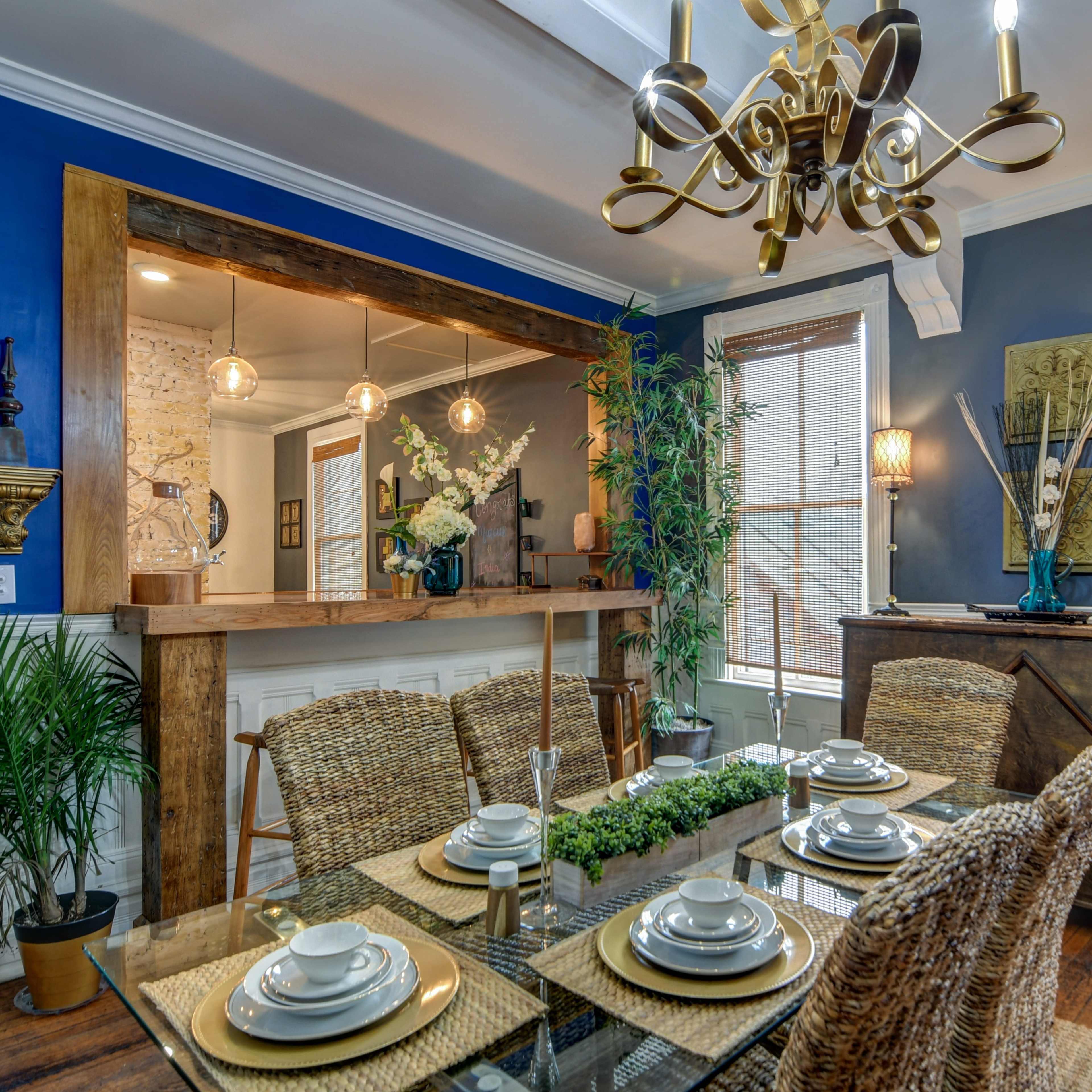 A dining room features a glass table set with plates and a centerpiece, surrounded by woven chairs, with blue walls and a wooden sideboard.