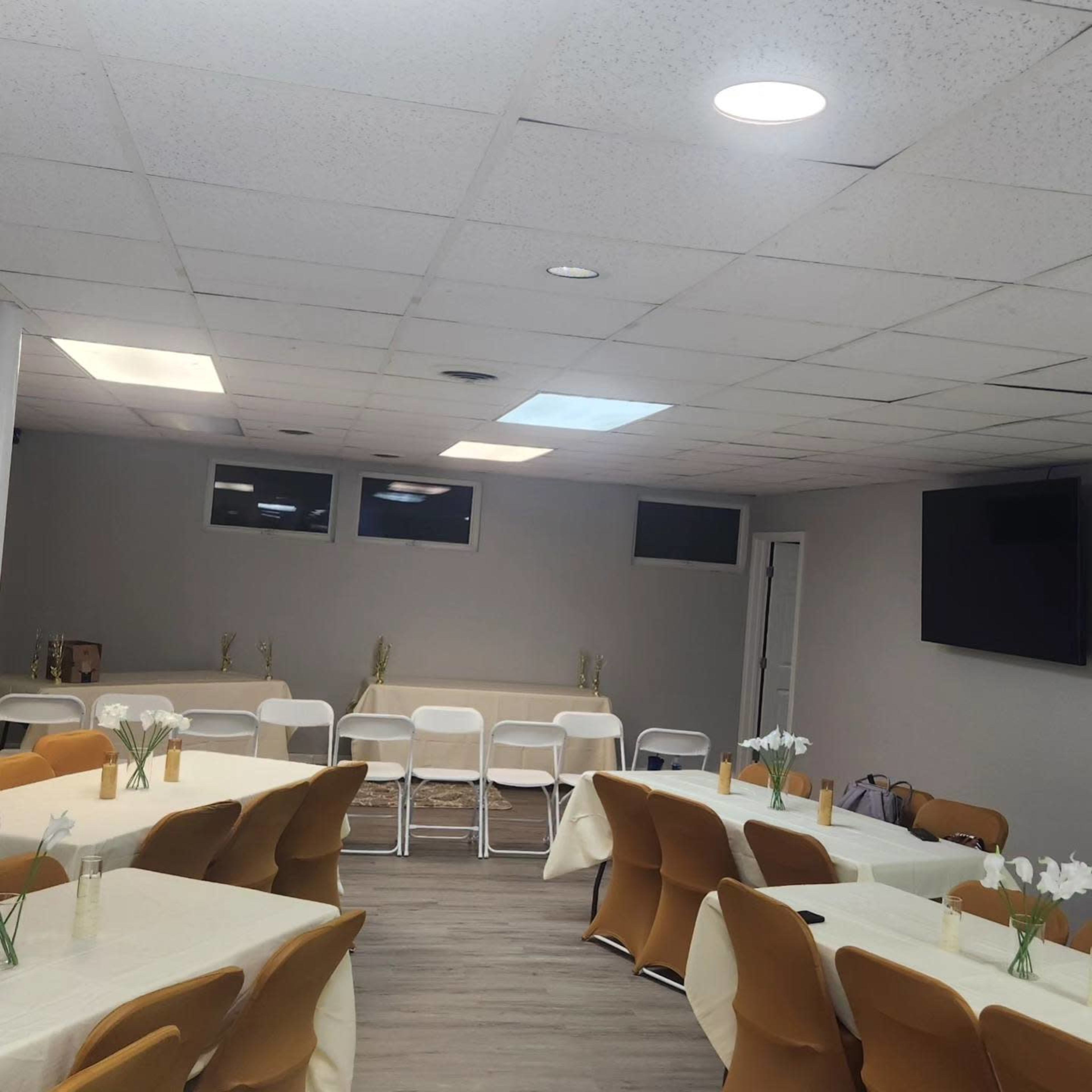A large, well-lit room features neatly arranged tables with white tablecloths and chairs, surrounded by windows and a wall-mounted television.