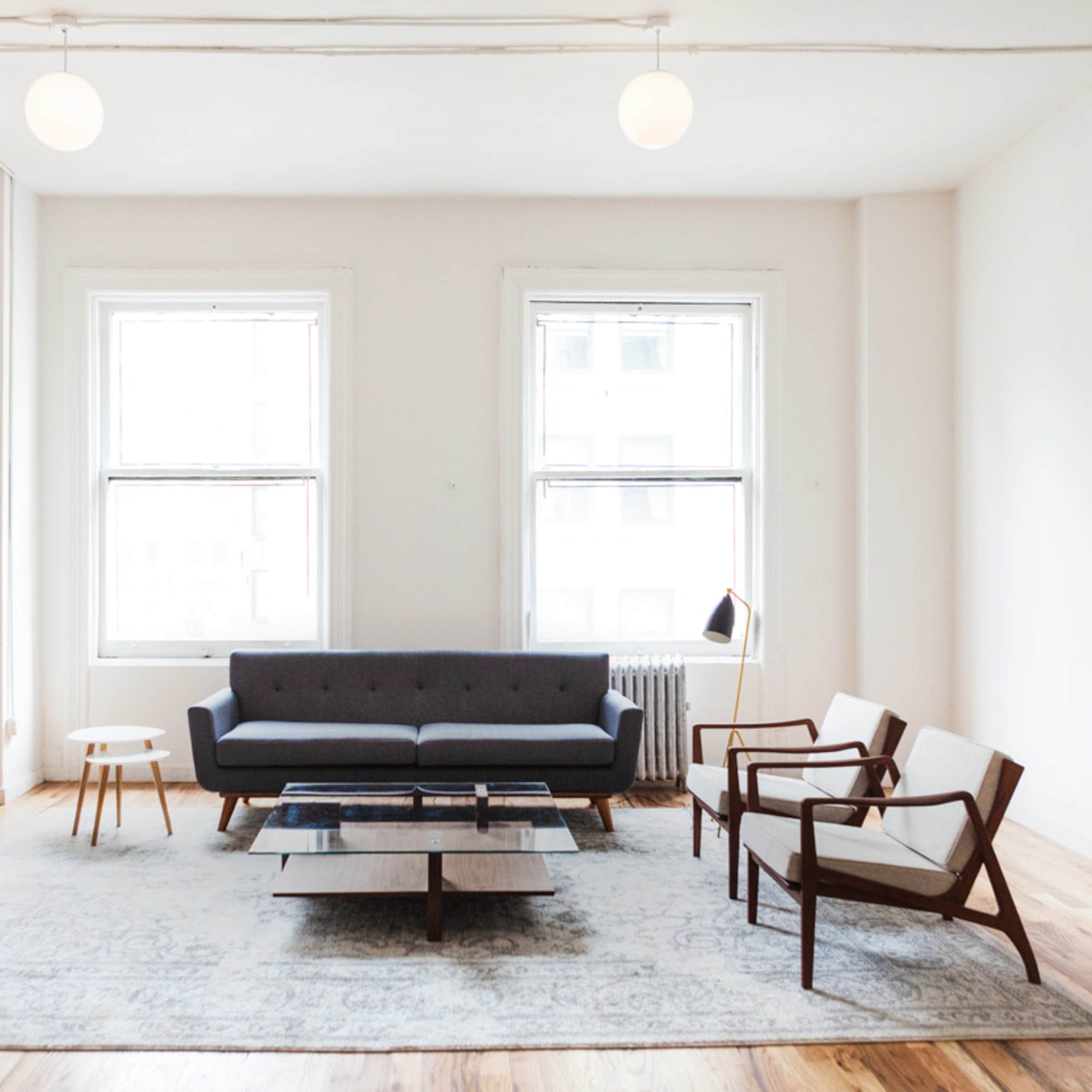 A minimalist living room features a gray sofa, two wooden armchairs, a coffee table, and large windows allowing natural light to fill the space.