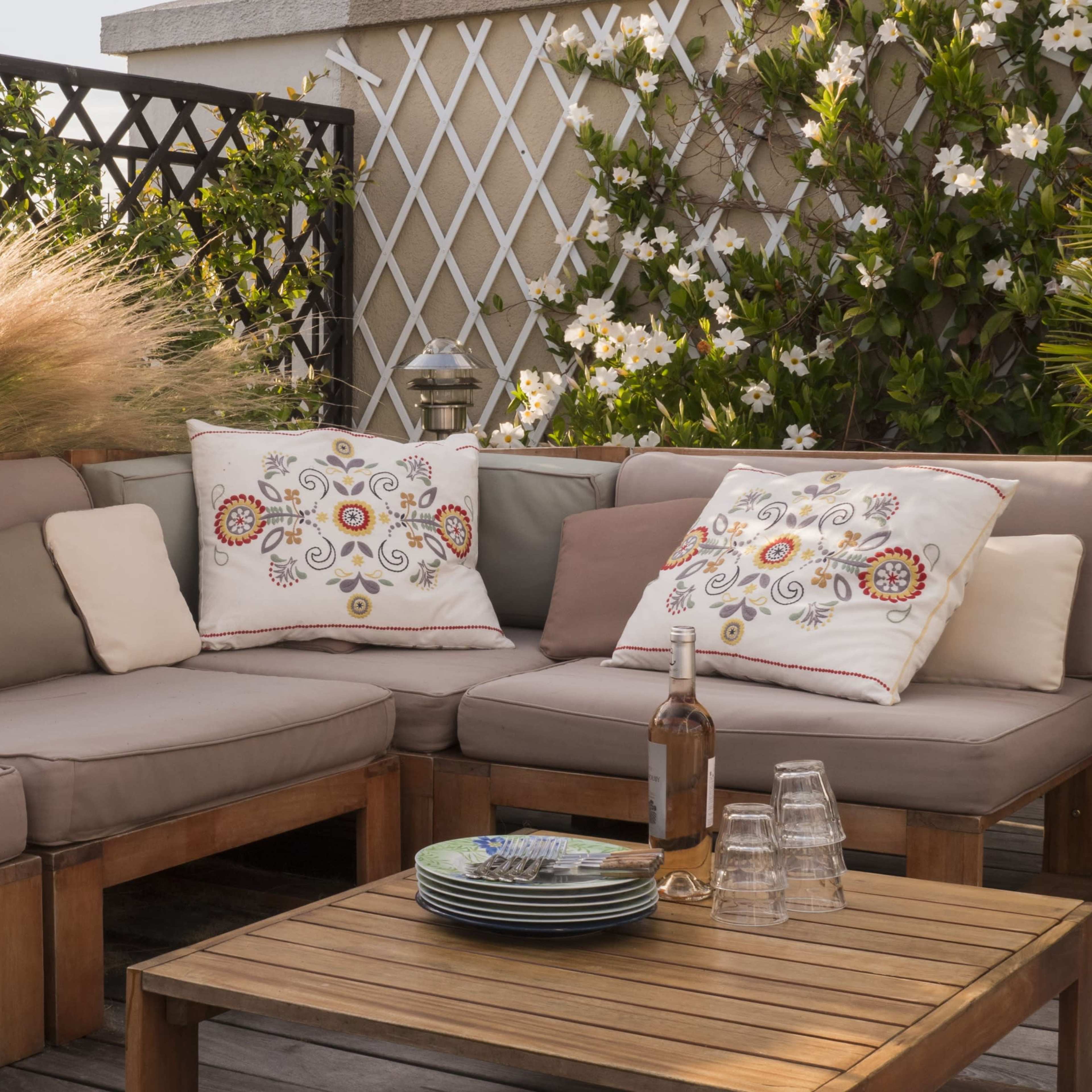 A comfortable outdoor seating area features a large sectional sofa with decorative pillows, a wooden coffee table, and greenery in the background.