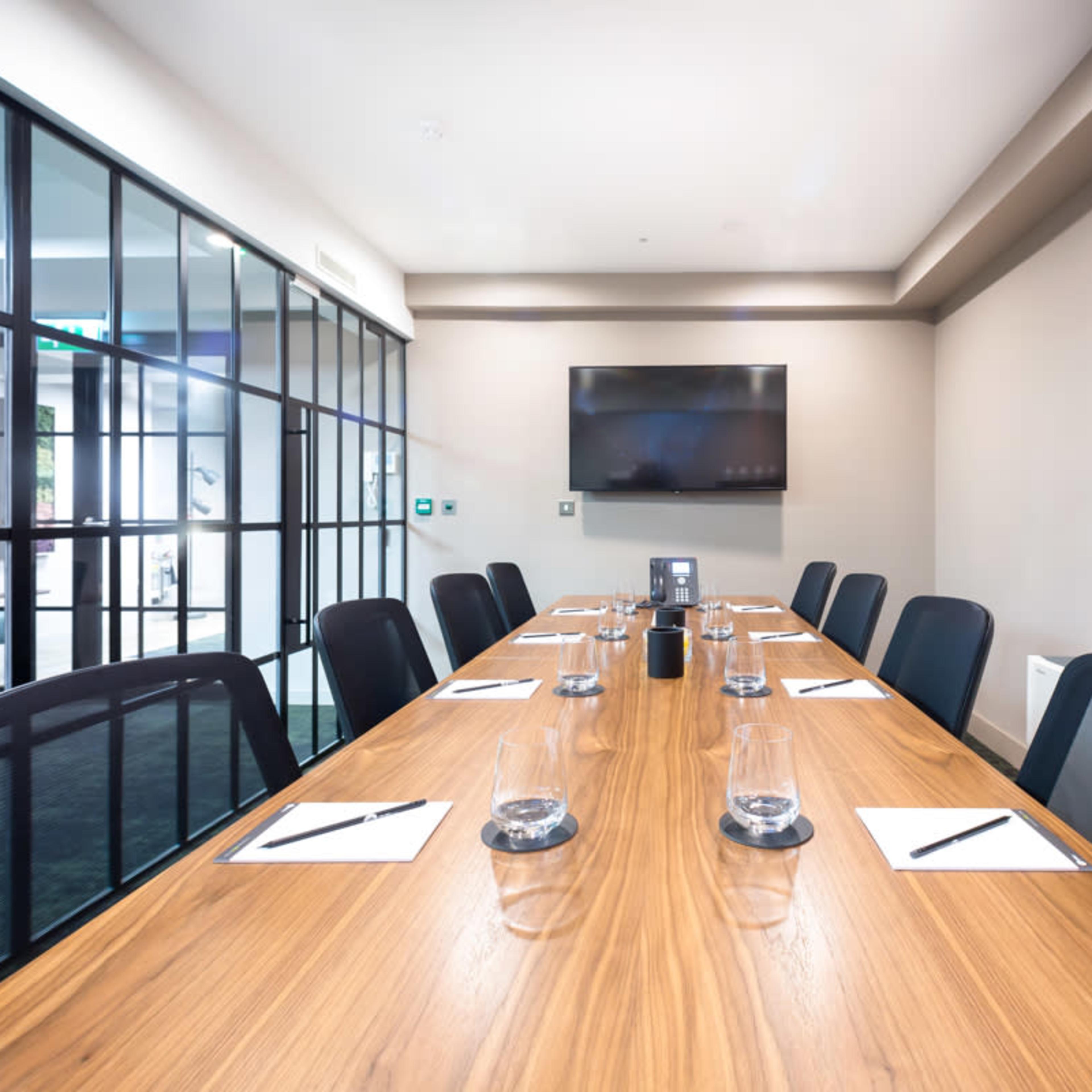 A conference room with a long wooden table set for a meeting, complete with notebooks, pens, and glasses of water, is displayed.