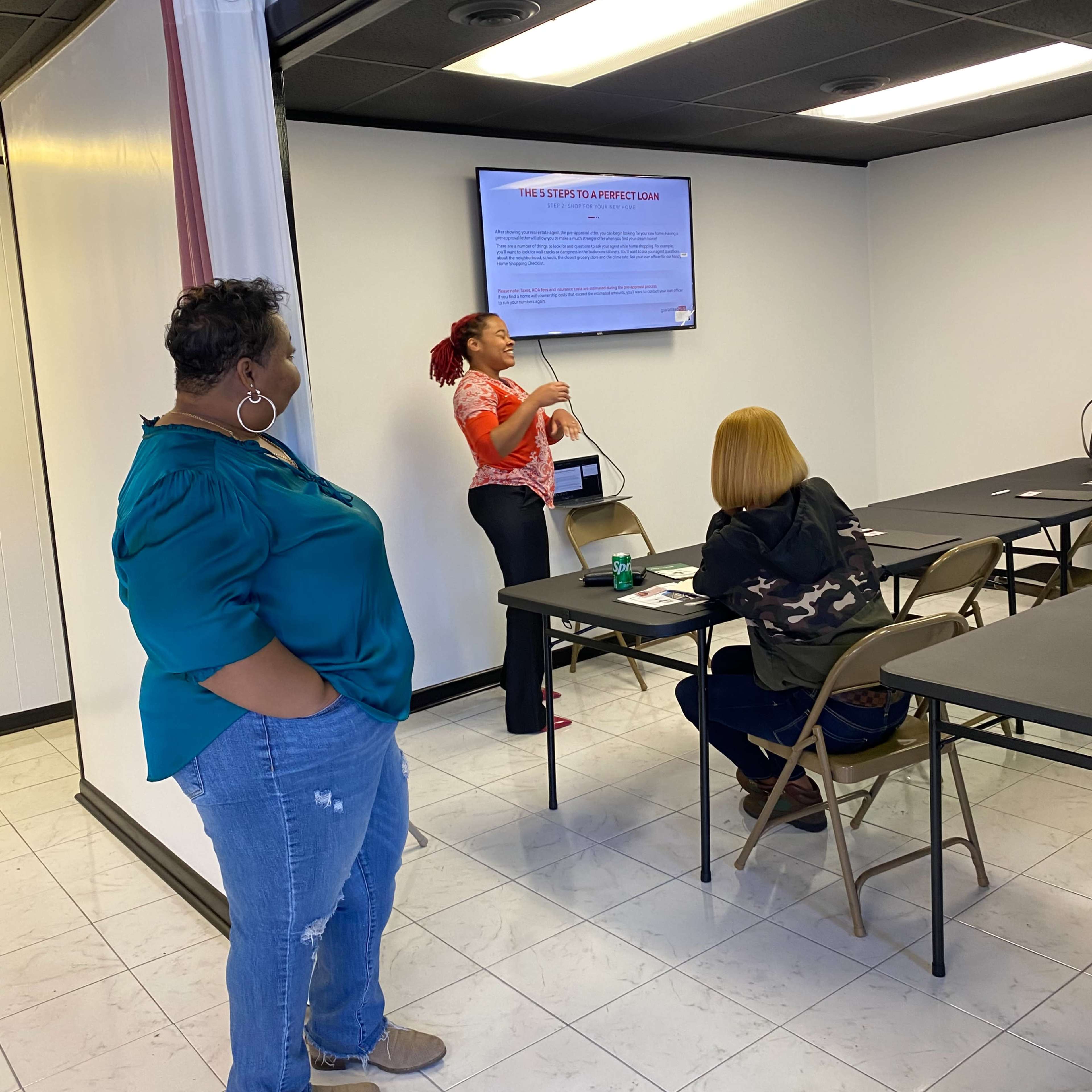 A woman stands at the front of a classroom, presenting information on a screen while two seated individuals listen attentively.