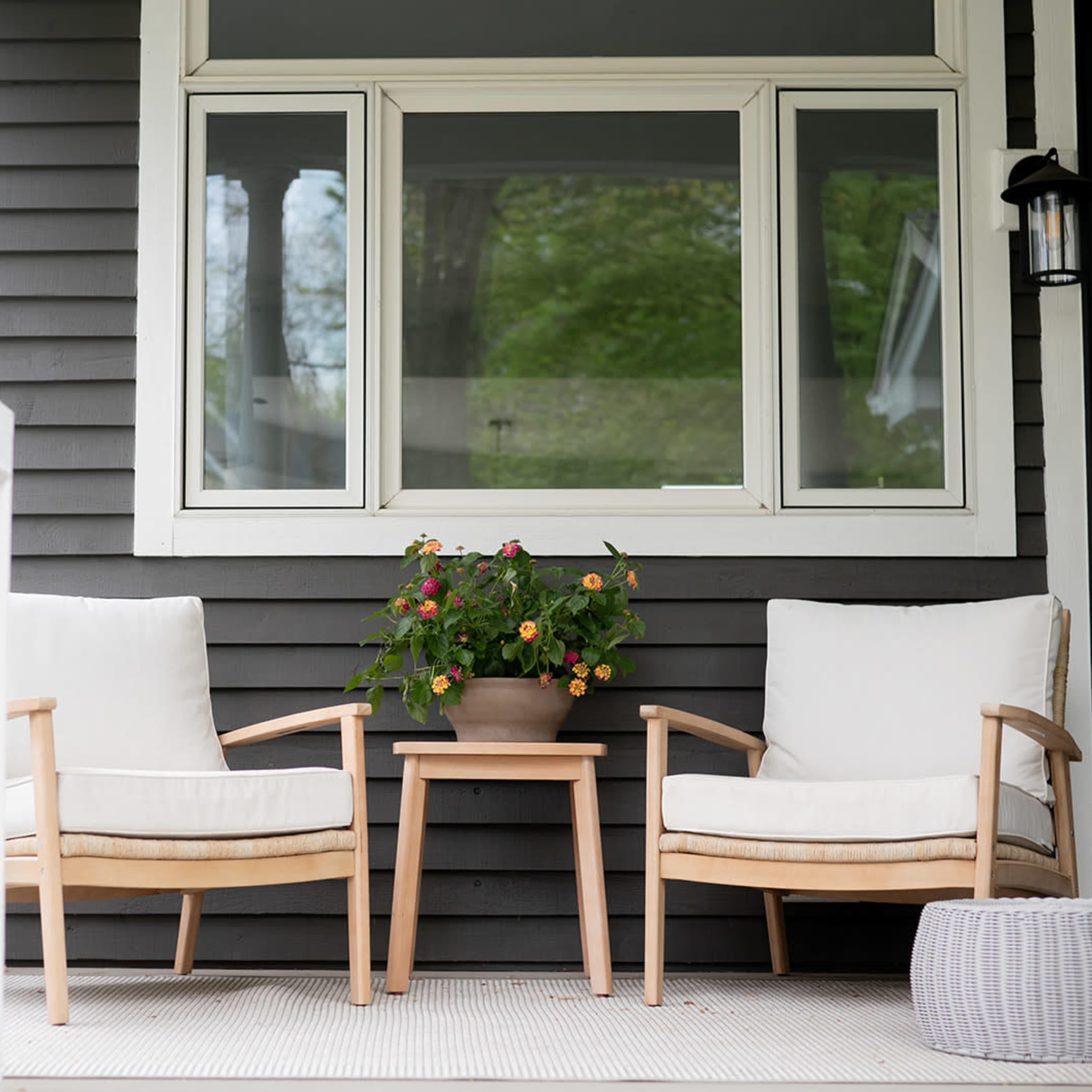 Two wooden chairs with white cushions and a small table sit on a porch beside a potted plant, with a view of a house's exterior and a window.