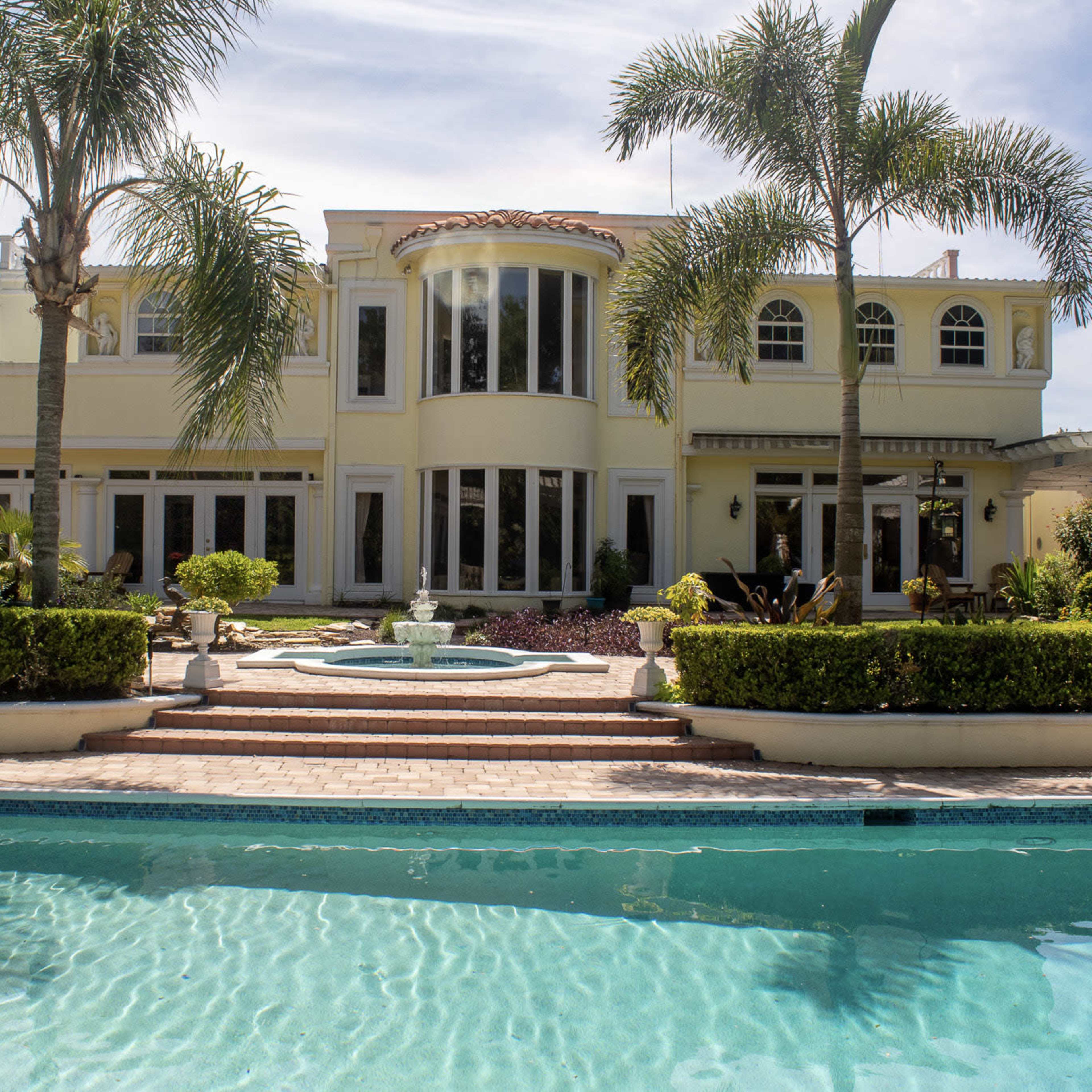 A large, modern house with multiple windows and a pool in the foreground, surrounded by neatly maintained landscaping and palm trees.