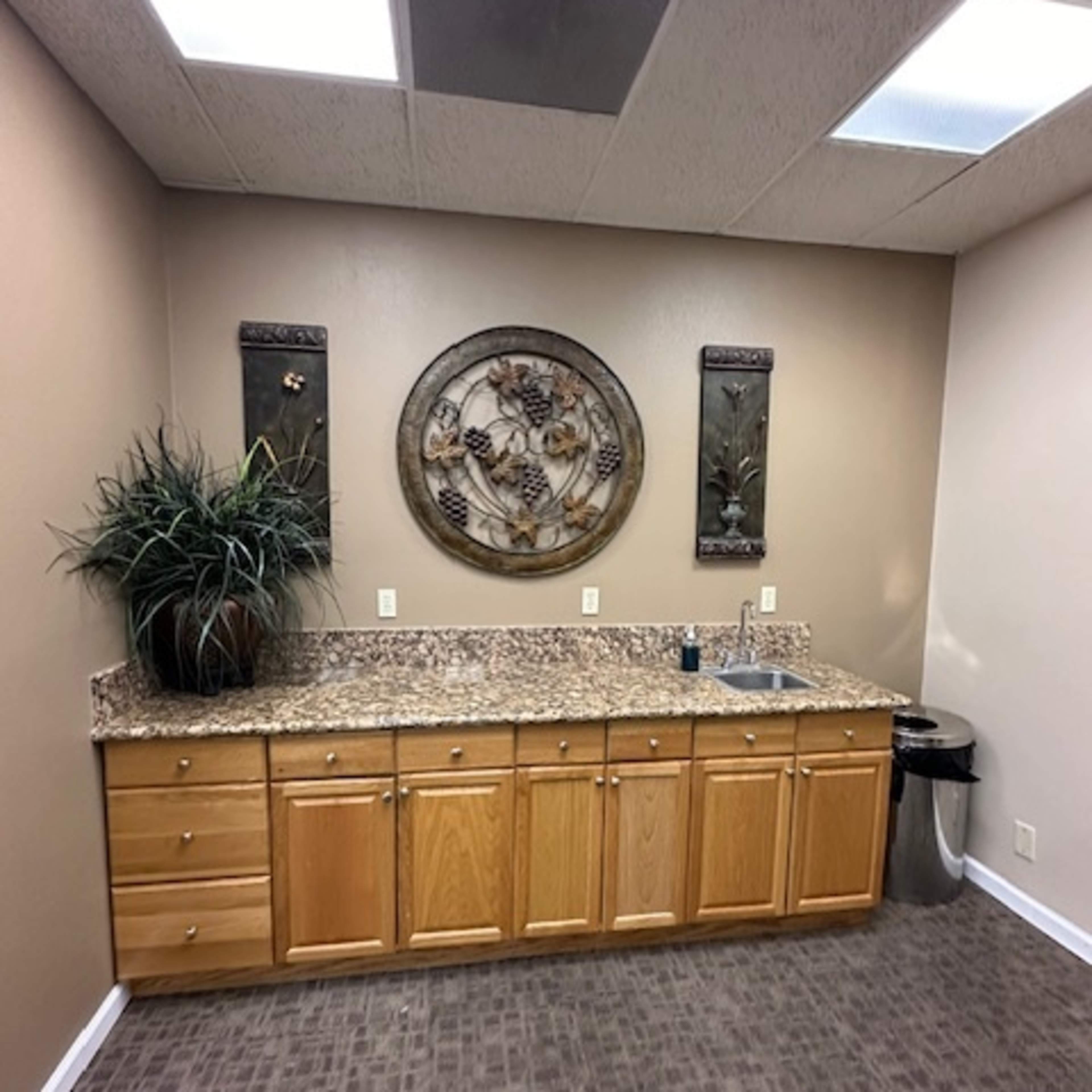 A small kitchenette with wooden cabinets, a granite countertop, a sink, and decorative wall art, set against a neutral-toned wall.
