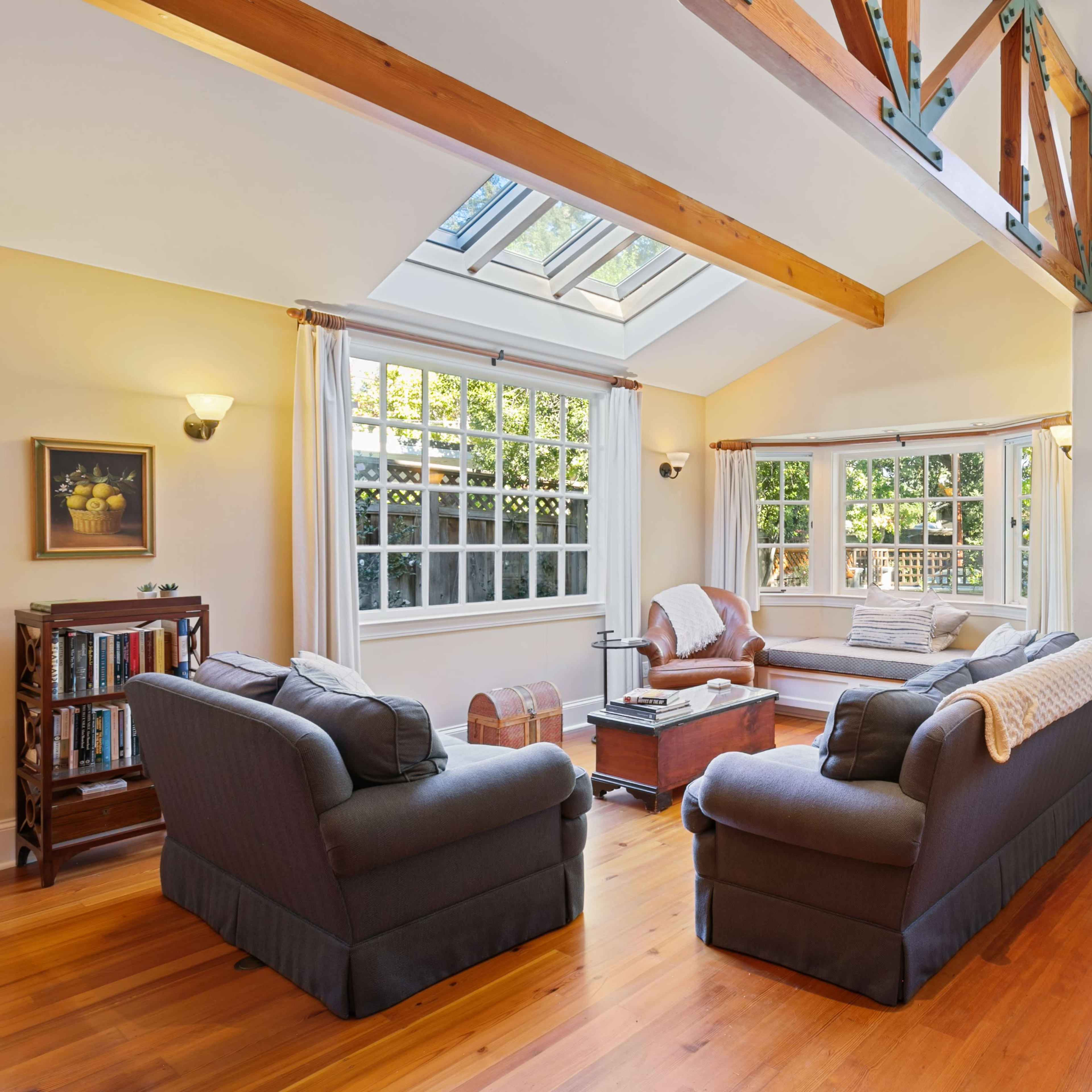 A sunlit living room with a high ceiling, featuring two sofas, a coffee table, and large windows that provide a view of the outdoors.