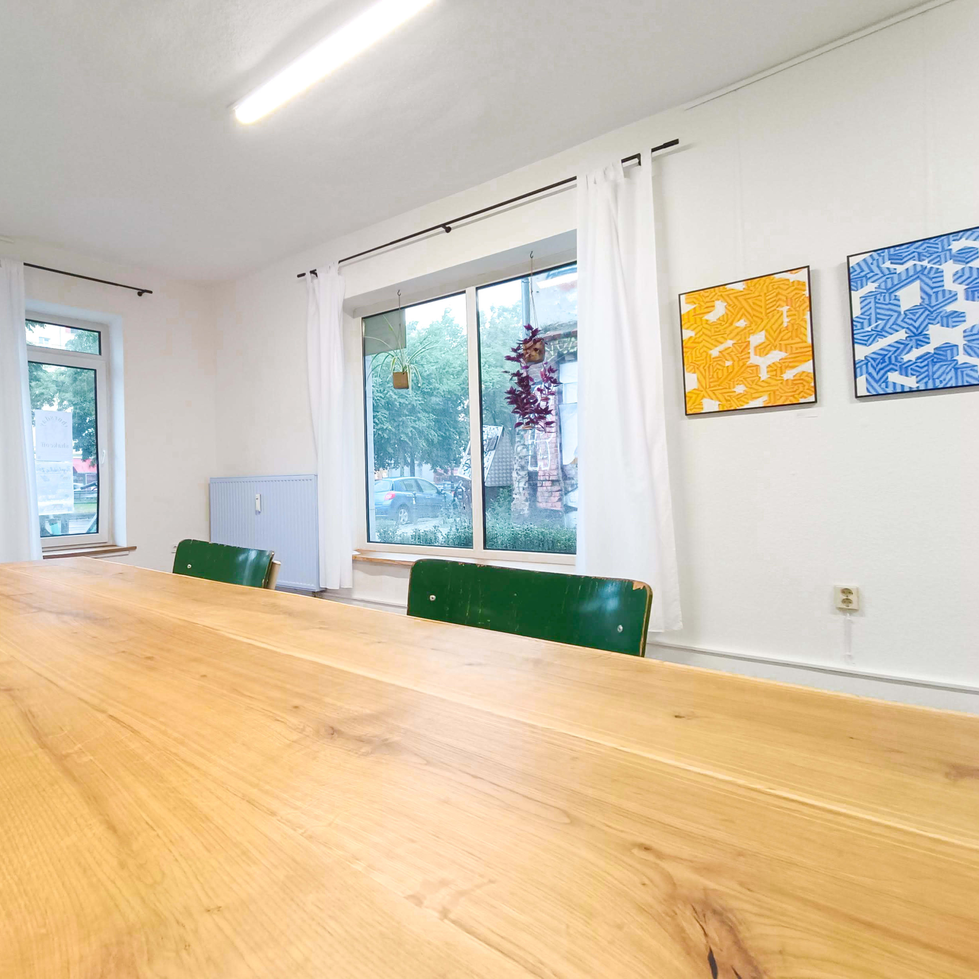 A wooden dining table with green chairs, set in a room featuring large windows and framed artwork on the walls.