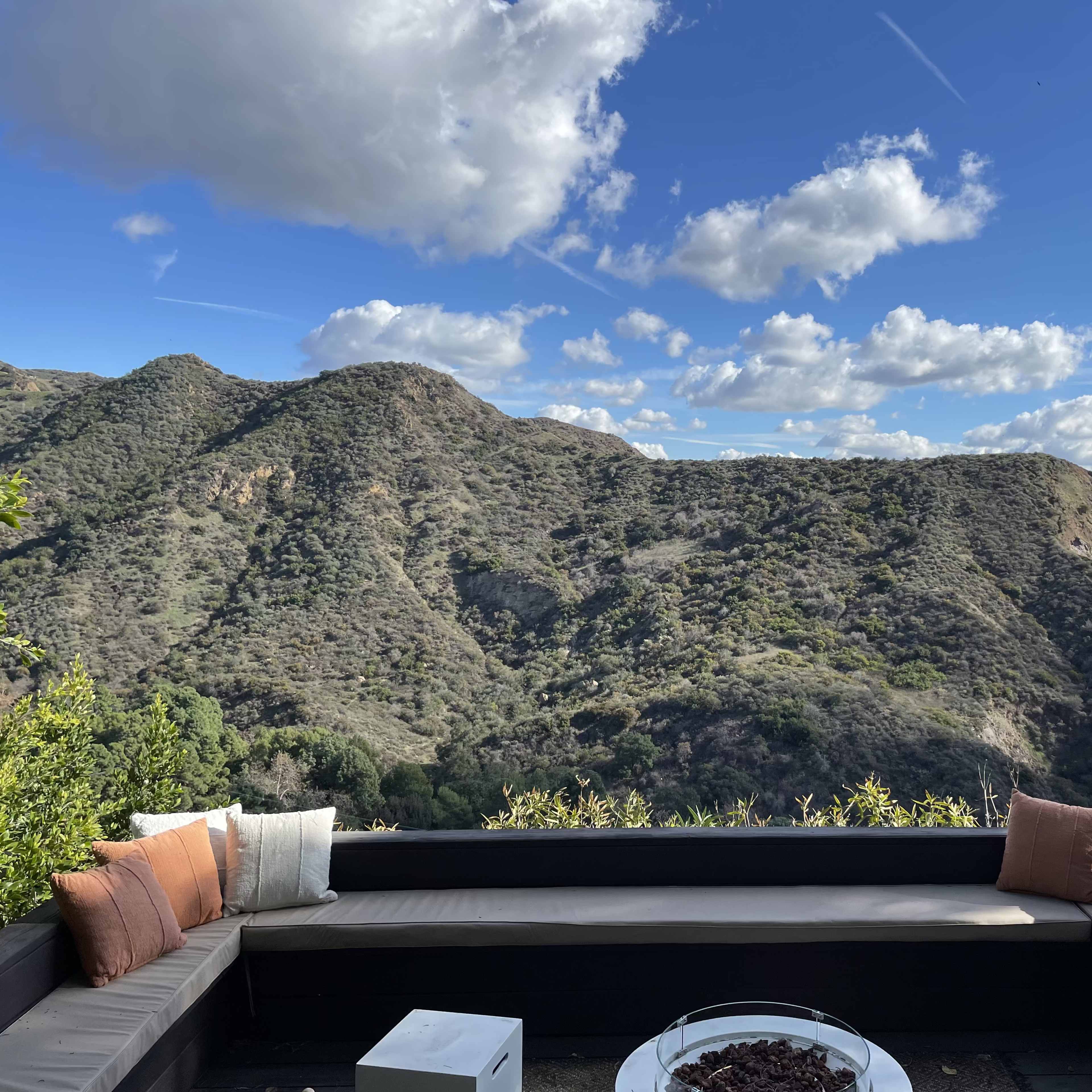 A seating area with cushions overlooking a hilly landscape under a partly cloudy sky.